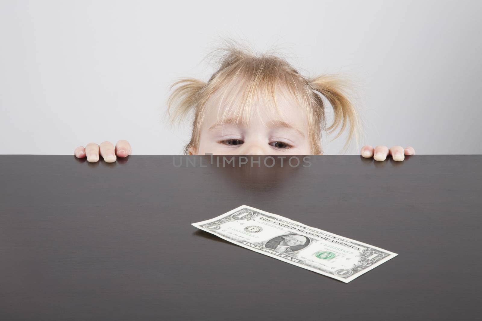 portrait of blonde caucasian baby nineteen month age with pigtails chubby face yellow shirt looking at dollar banknote on brown table horizontal