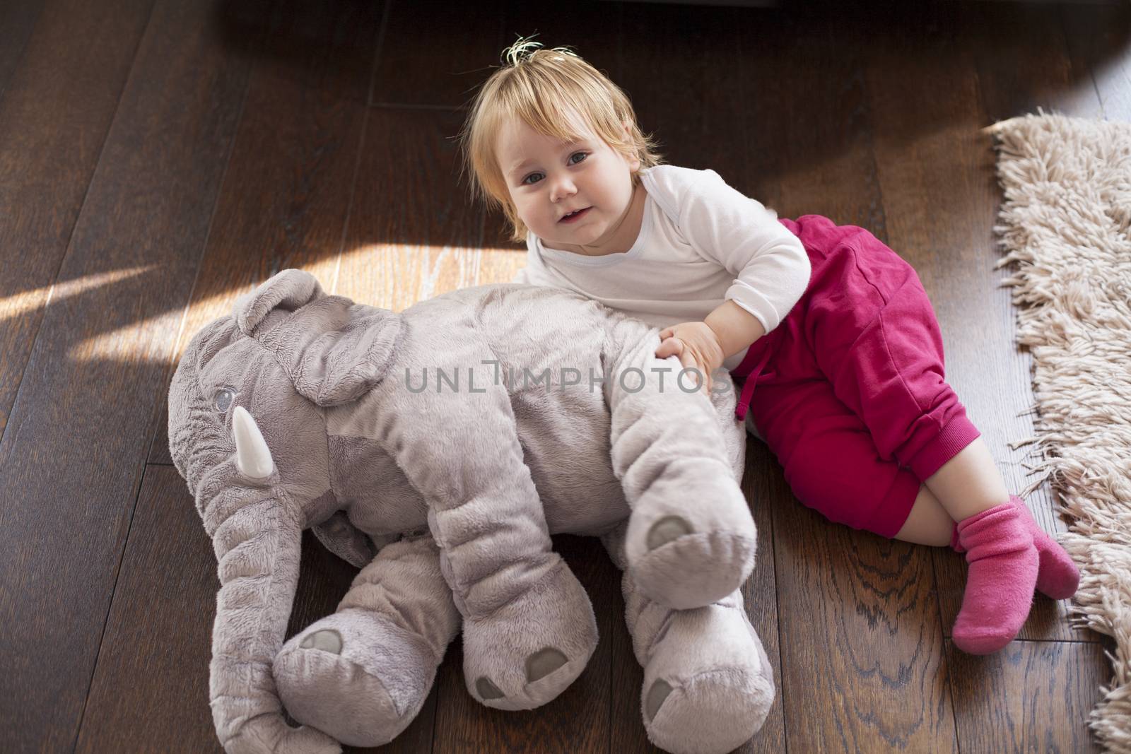portrait of blonde caucasian baby nineteen month age looking at camera smiling with cheerful expression face together grey plush doll on brown wooden floor