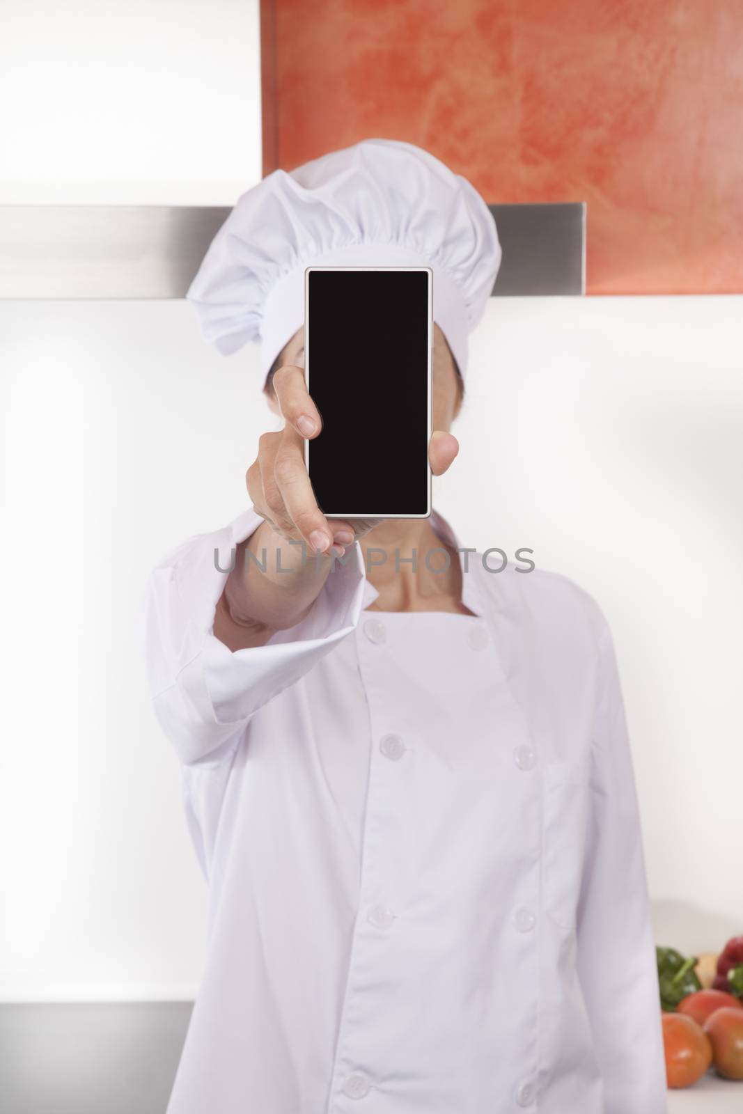 portrait of brunette happy chef woman with professional jacket and hat in white and orange kitchen showing blank screen mobile phone