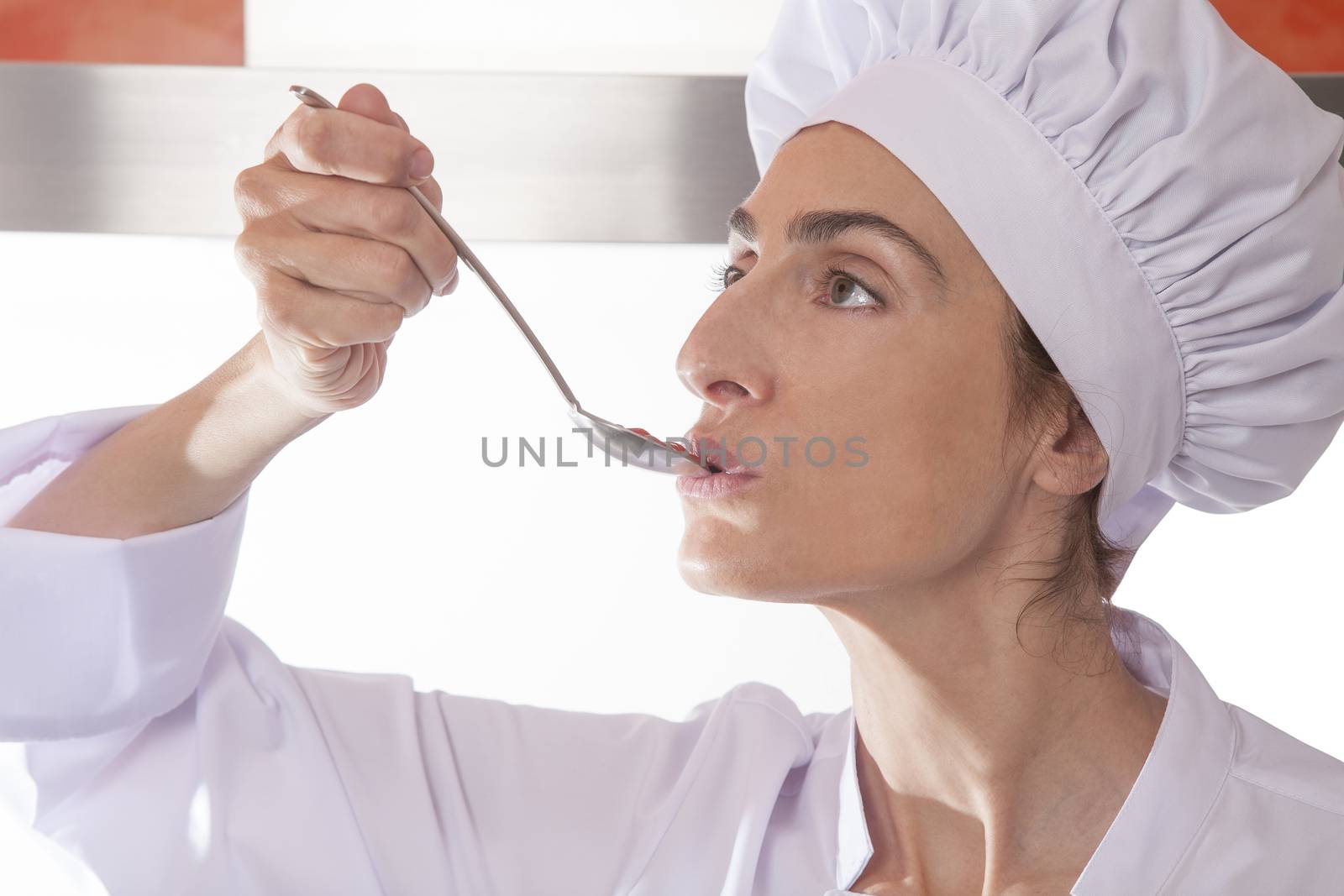 portrait of chef woman with professional jacket and hat tasting red sauce or cream puree like tomato ketchup in a steel soup spoon in her hand