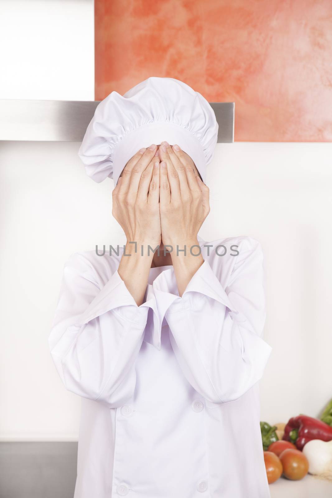 portrait of brunette chef woman with professional jacket and hat in white and orange kitchen hidden face with both hands