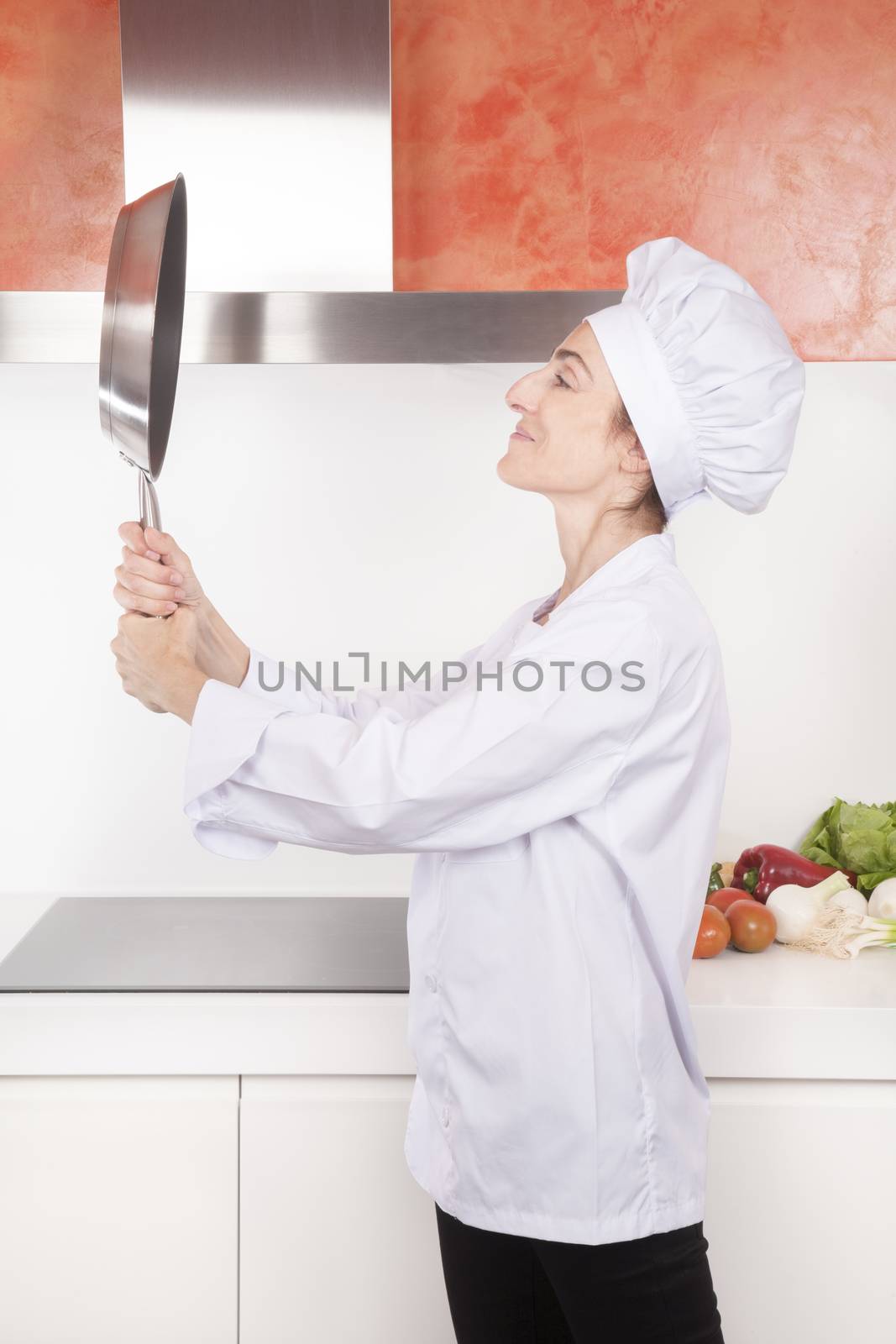 portrait of happy brunette chef woman with professional jacket and hat in white and orange kitchen looking herself at steel blank pan like a mirror