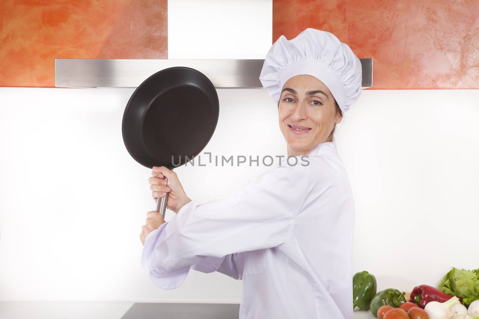 portrait of happy brunette chef woman with professional jacket and hat in white and orange kitchen ready to strike with steel blank pan