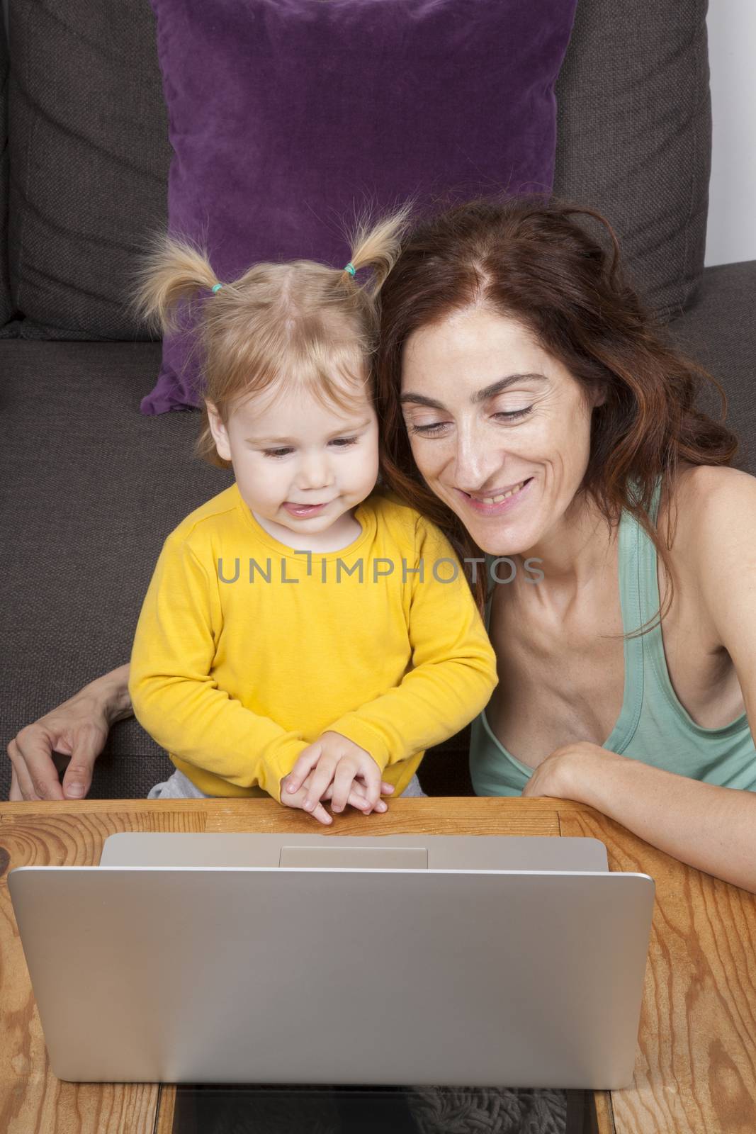 nineteen month aged blonde baby sticking her tongue out with pigtails and brunette woman mother looking at grey laptop on brown table