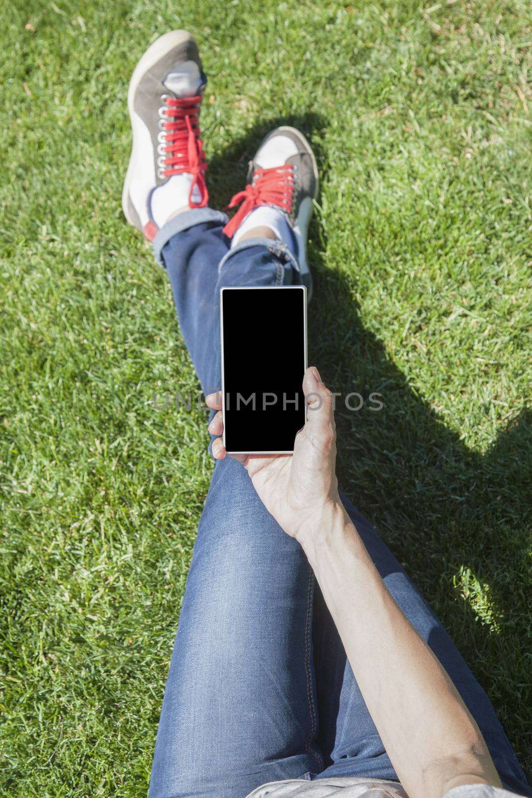 hand of woman blue jeans trousers legged with mobile phone smartphone blank screen sitting on green grass lawn in park