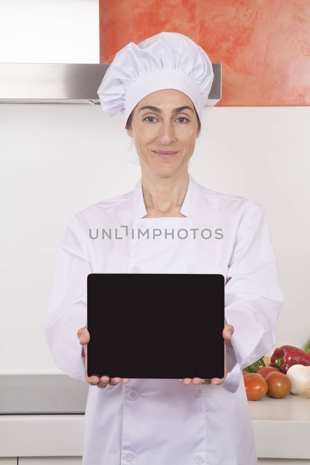 portrait of brunette happy chef woman with professional jacket and hat in white and orange kitchen showing blank screen tablet horizontal