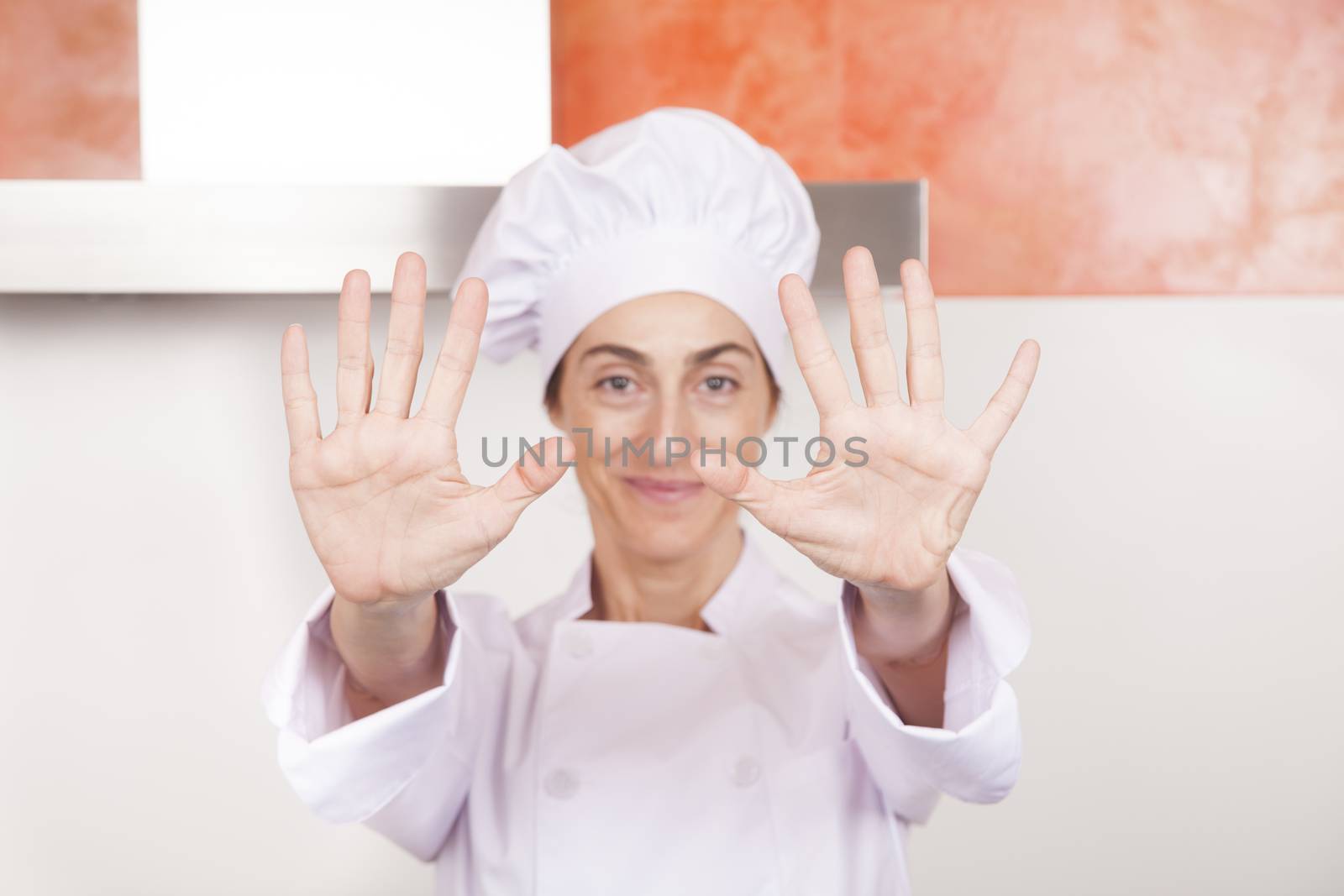 portrait of brunette happy chef woman with professional jacket and hat in white and orange kitchen showing both open palms of hands