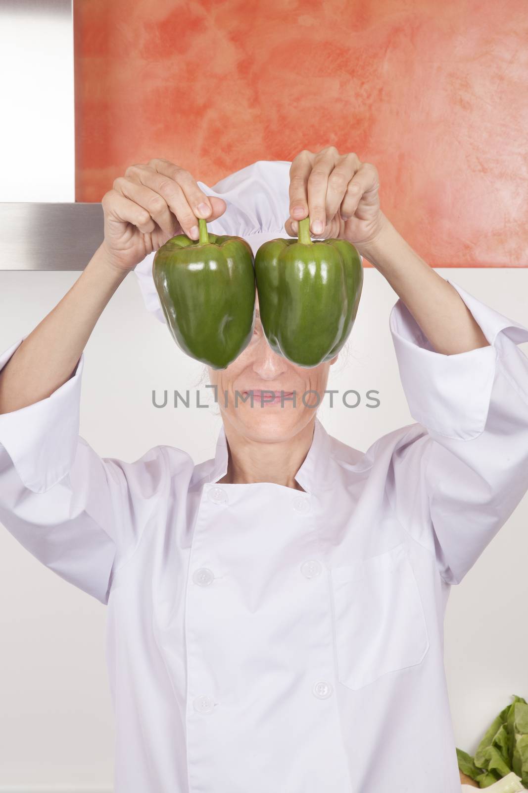 portrait of brunette happy chef woman with professional jacket and hat  in white and orange kitchen with two raw fresh big green peppers in her hands