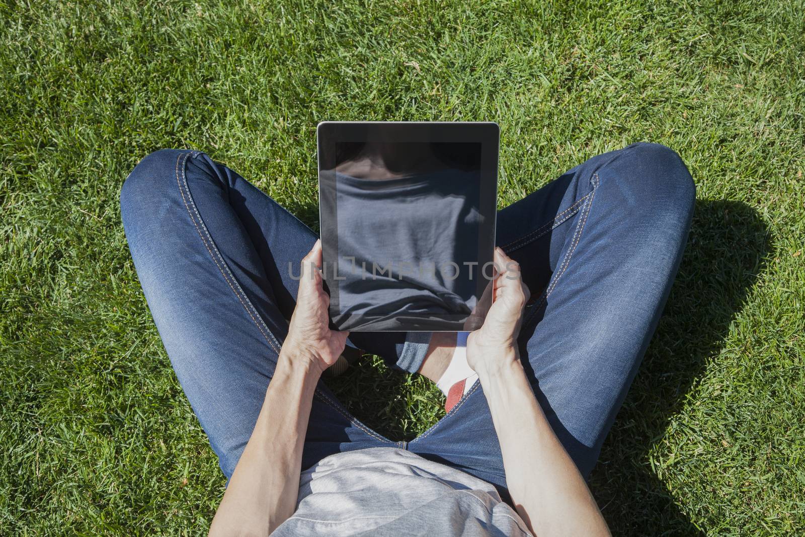 woman blue jeans trousers grey shirt legged with digital tablet blank screen in her hands sitting on green grass lawn in park