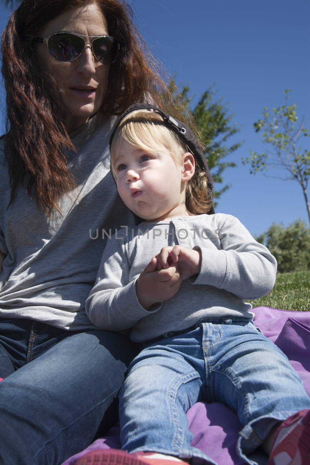 portrait of two years age blonde baby with black cap eating banana next to woman mother in park