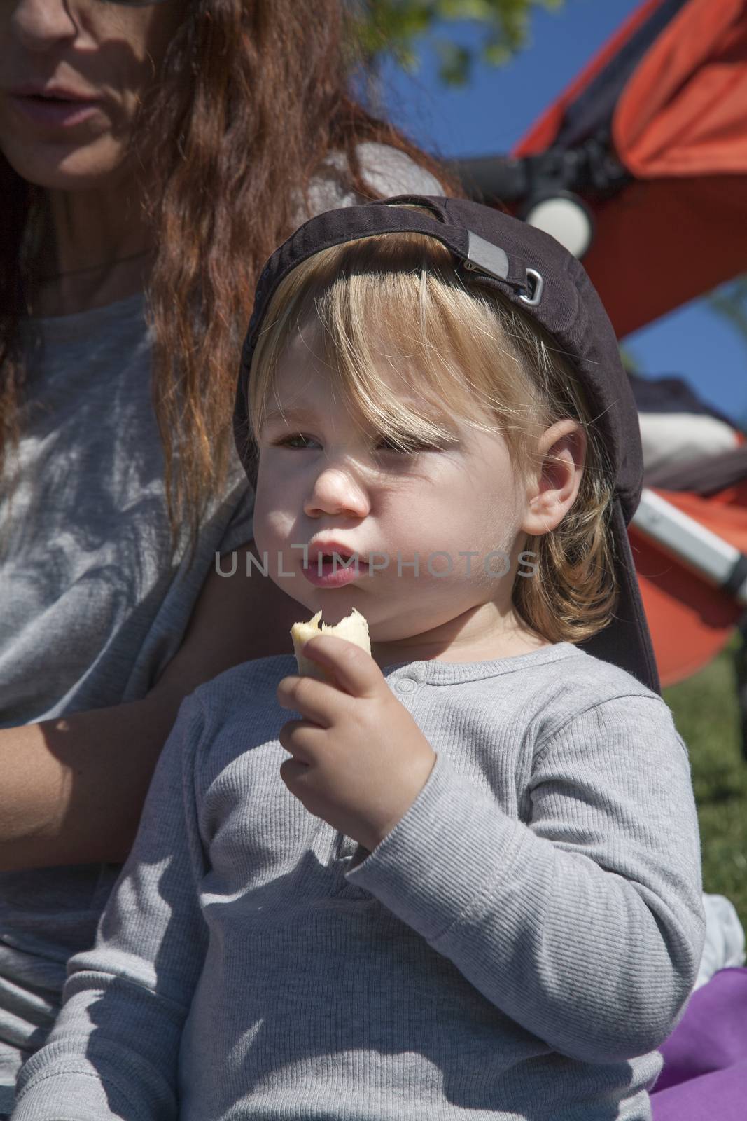 portrait of two years age blonde baby with black cap eating banana next to woman mother