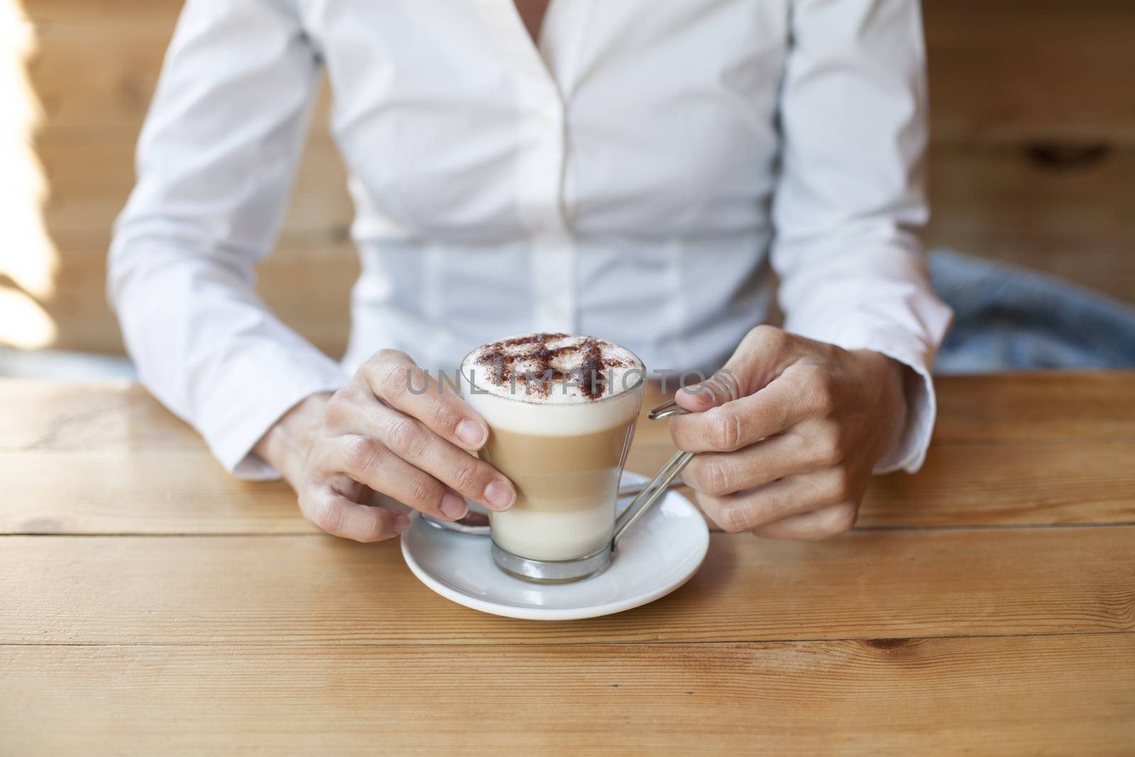 cappuccino coffee cup in hands of woman with white shirt on light brown wooden table cafe