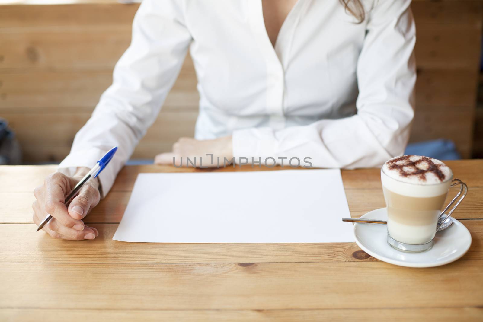 woman with white shirt and cappuccino coffee cup ready to write on white sheet paper on light brown wooden table