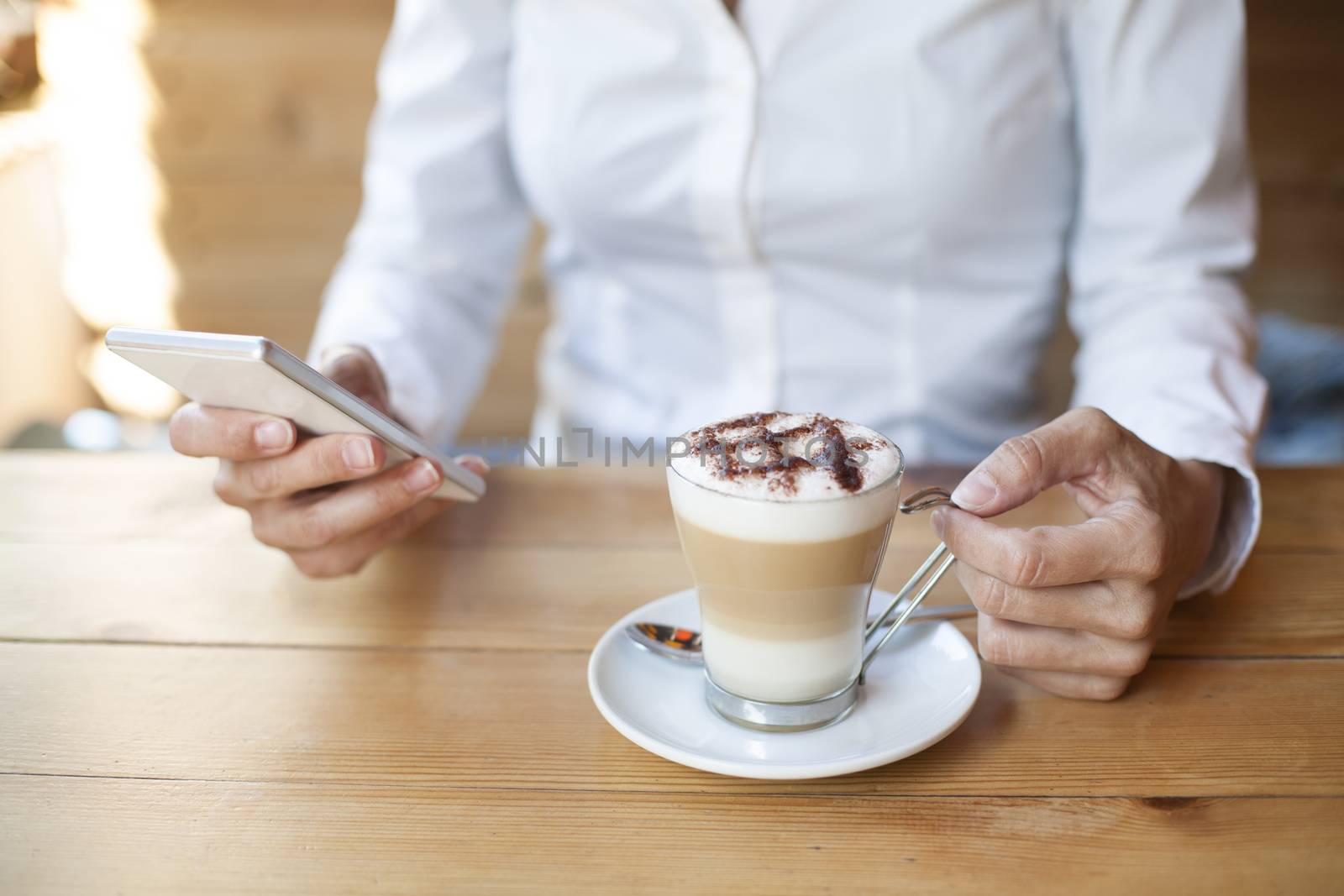 woman touching mobile phone with cappuccino coffee cup on light brown wooden table cafe
