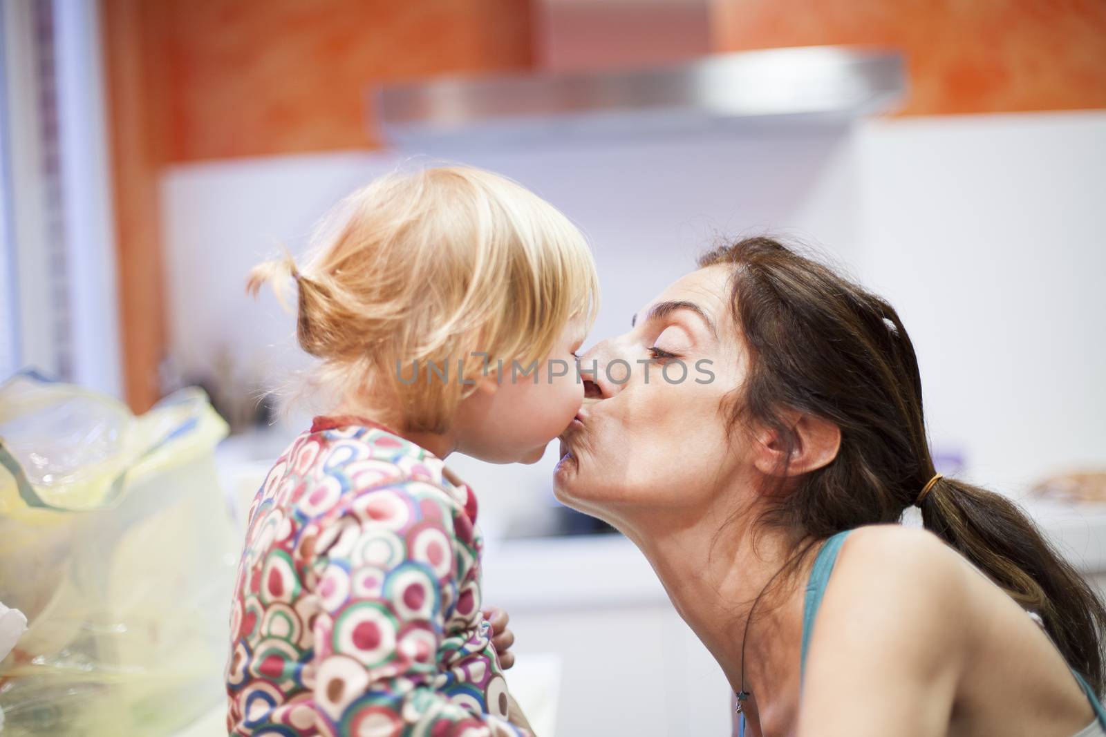 nineteen month aged blonde baby colored shirt with pigtails and brunette woman mother kissing in mouth in orange kitchen