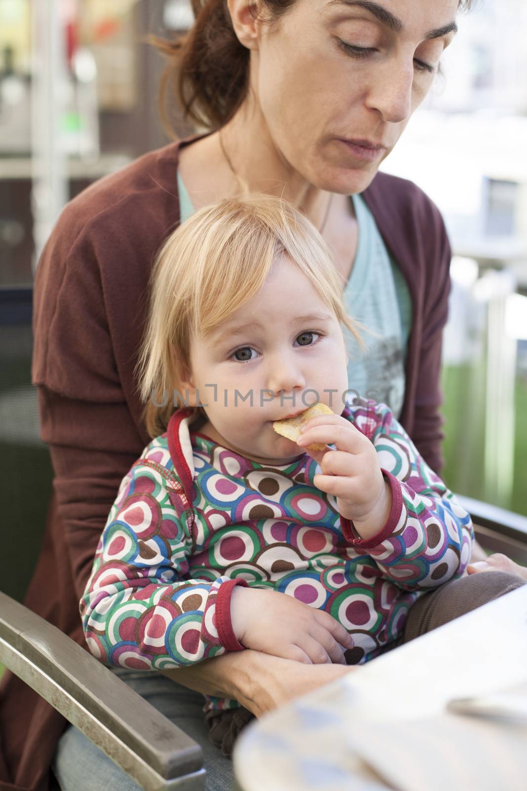 portrait of two years age blonde baby with colored shirt eating chips and looking at camera sitting on woman mother legs in table at cafe restaurant