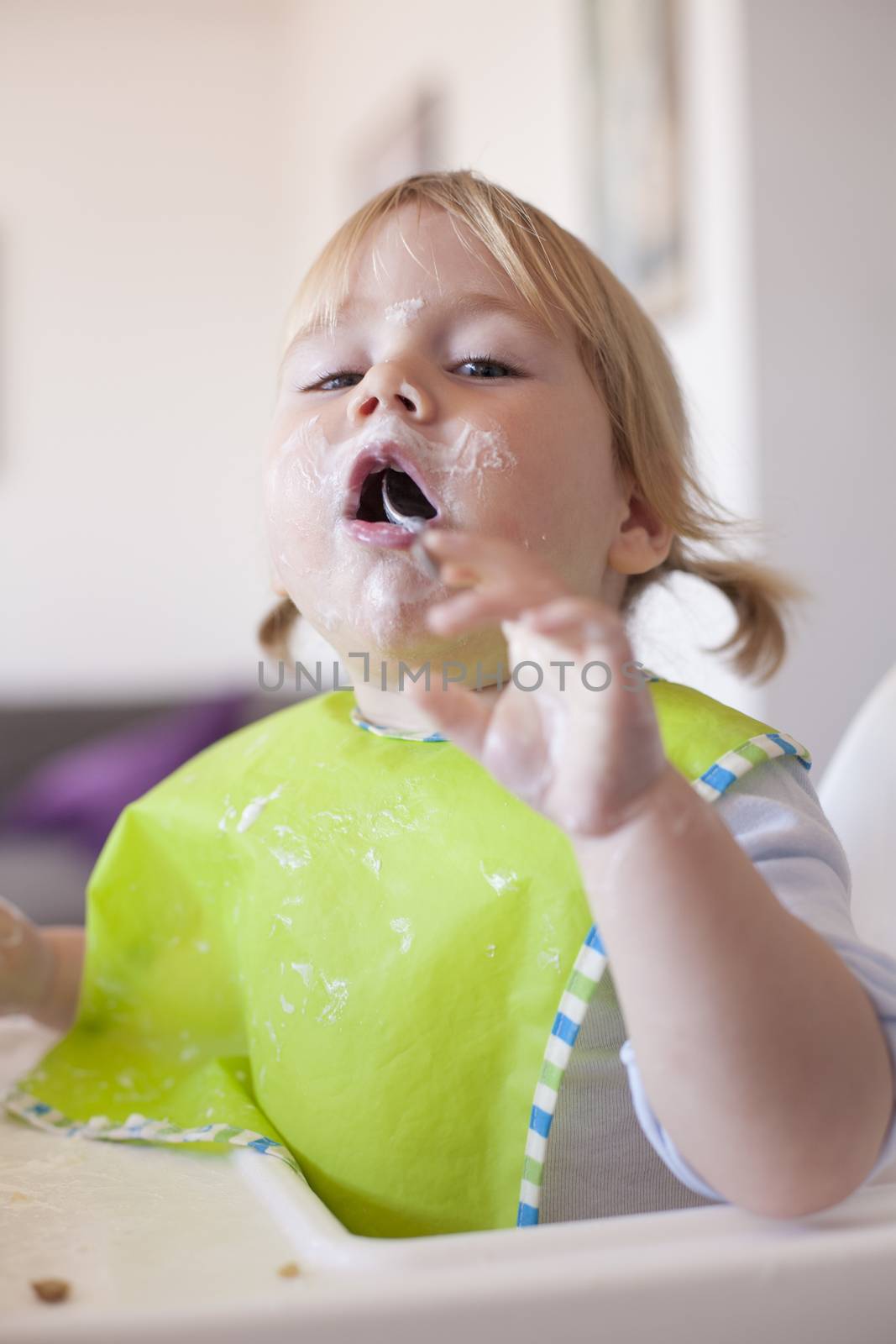 portrait of blonde caucasian baby nineteen month age chubby face looking at camera with green bib spoon in her mouth and smeared with yogurt