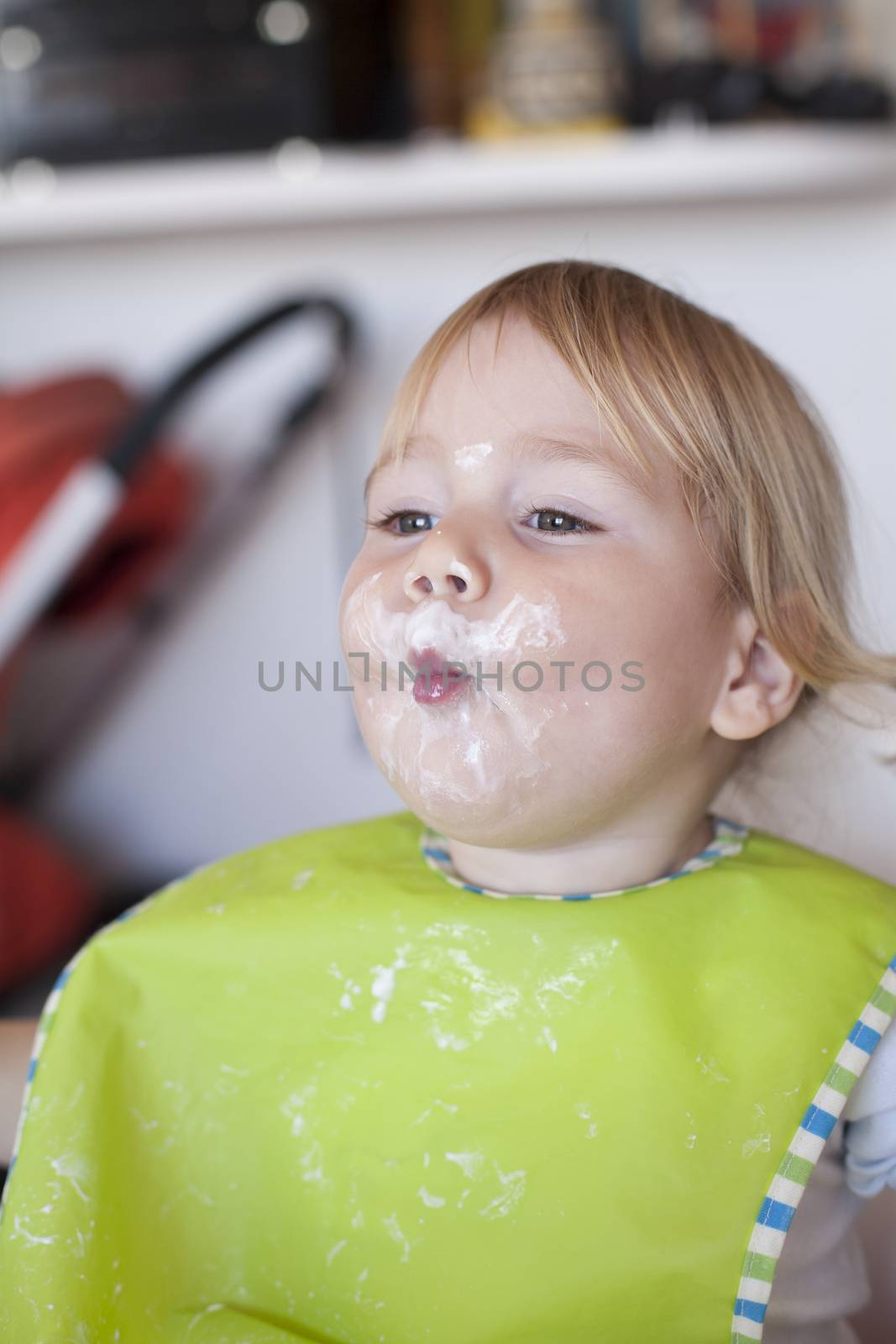 portrait of blonde caucasian baby nineteen month age chubby face looking with green bib sucked with mouth and smeared with yogurt