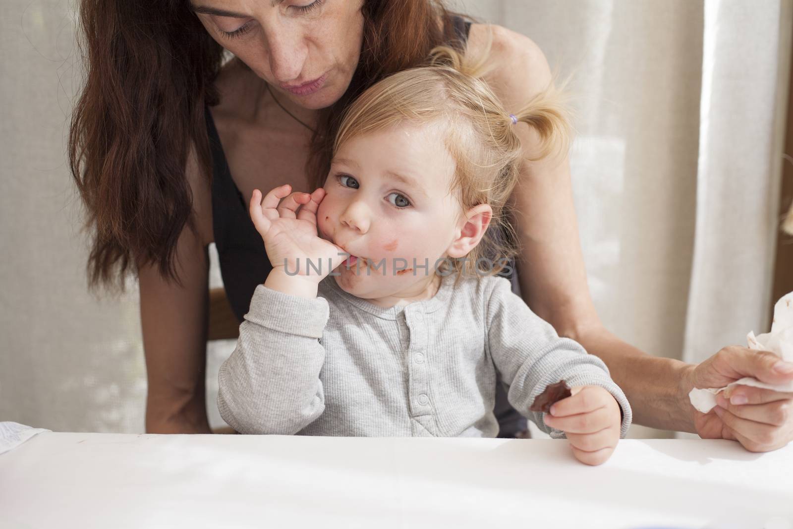 portrait of two years age blonde baby with grey shirt sucking her thumb finger eating chocolate piece sitting on legs of mother woman black shirt in white table
