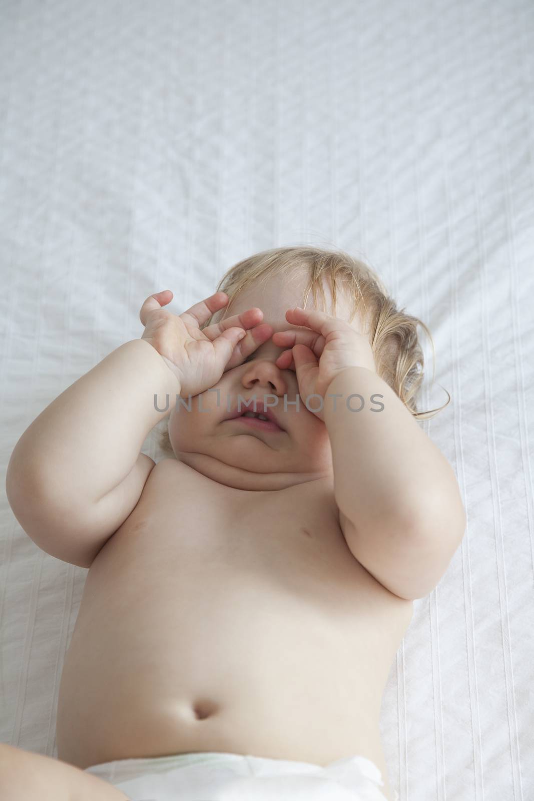 one year blonde baby diaper touching her eyes over white bedcover