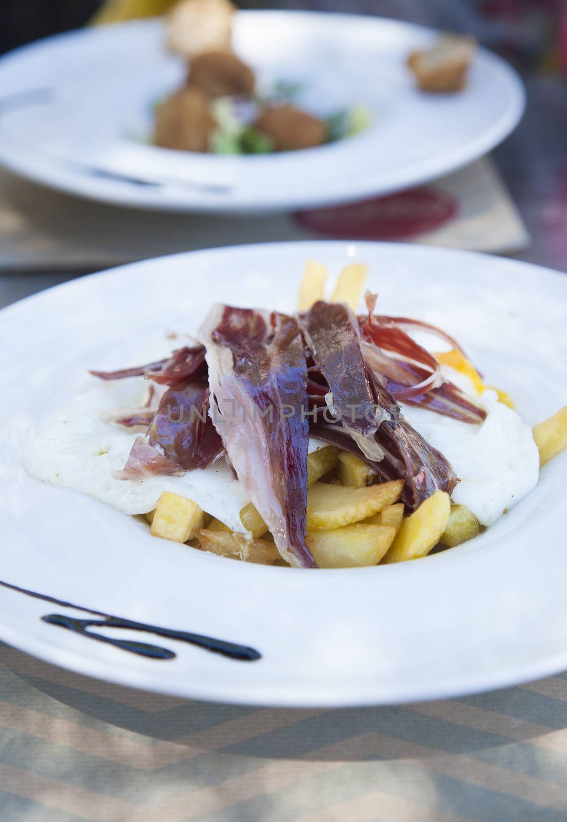 still life of white dish with typical Spain food named huevos rotos or broken eggs with potatoes french fries fried eggs and slices of iberico ham on table at cafe restaurant