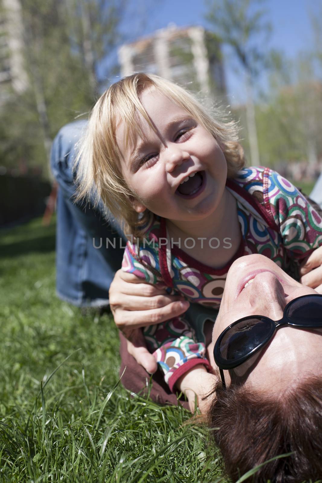 two years aged blonde happy baby colored shirt hugging with brunette woman mother with black sunglasses lying on green grass lawn