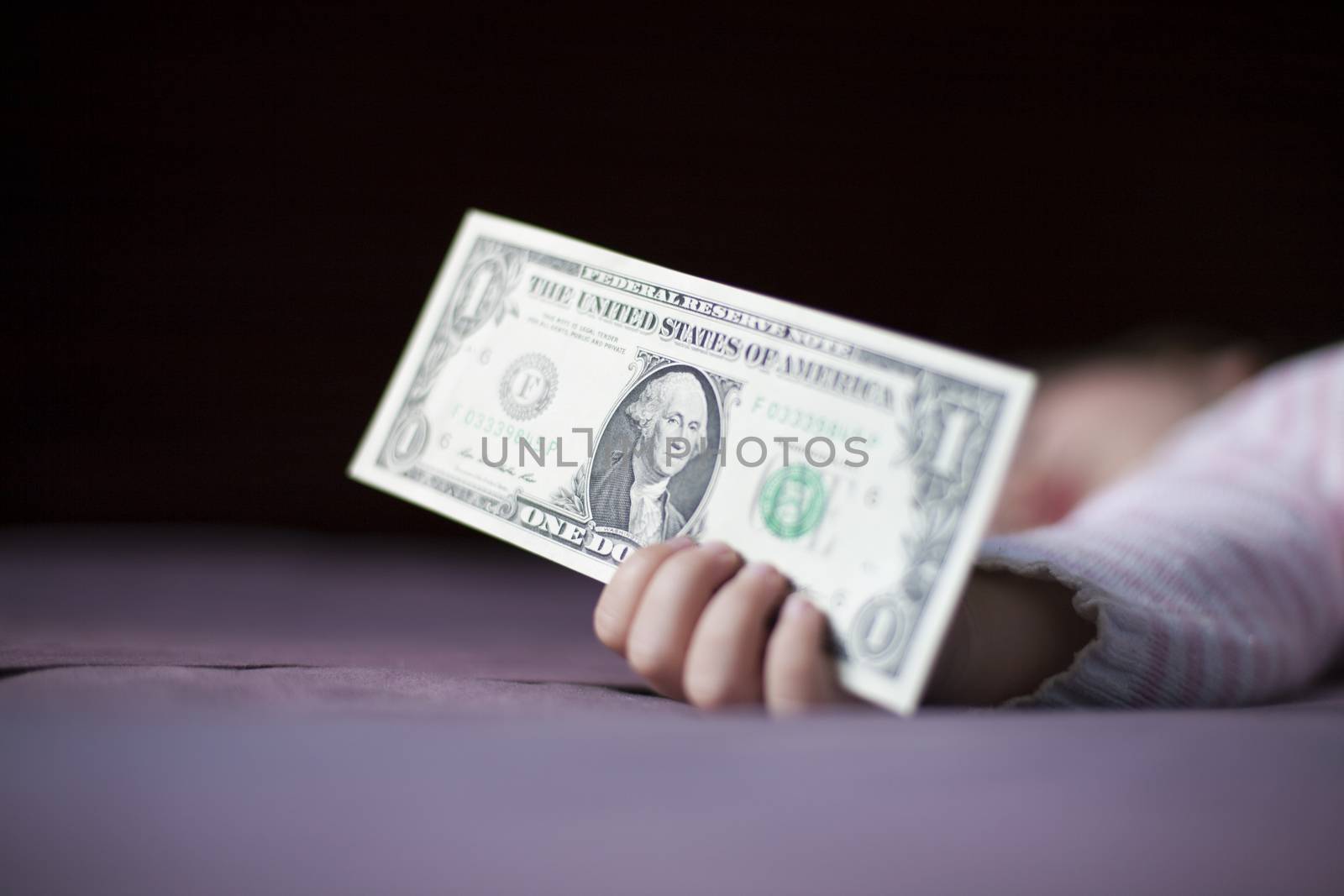 closeup one dollar banknote in hand of blonde caucasian baby nineteen month age with pink and white stripped jersey sleeping on brown sheets king bed