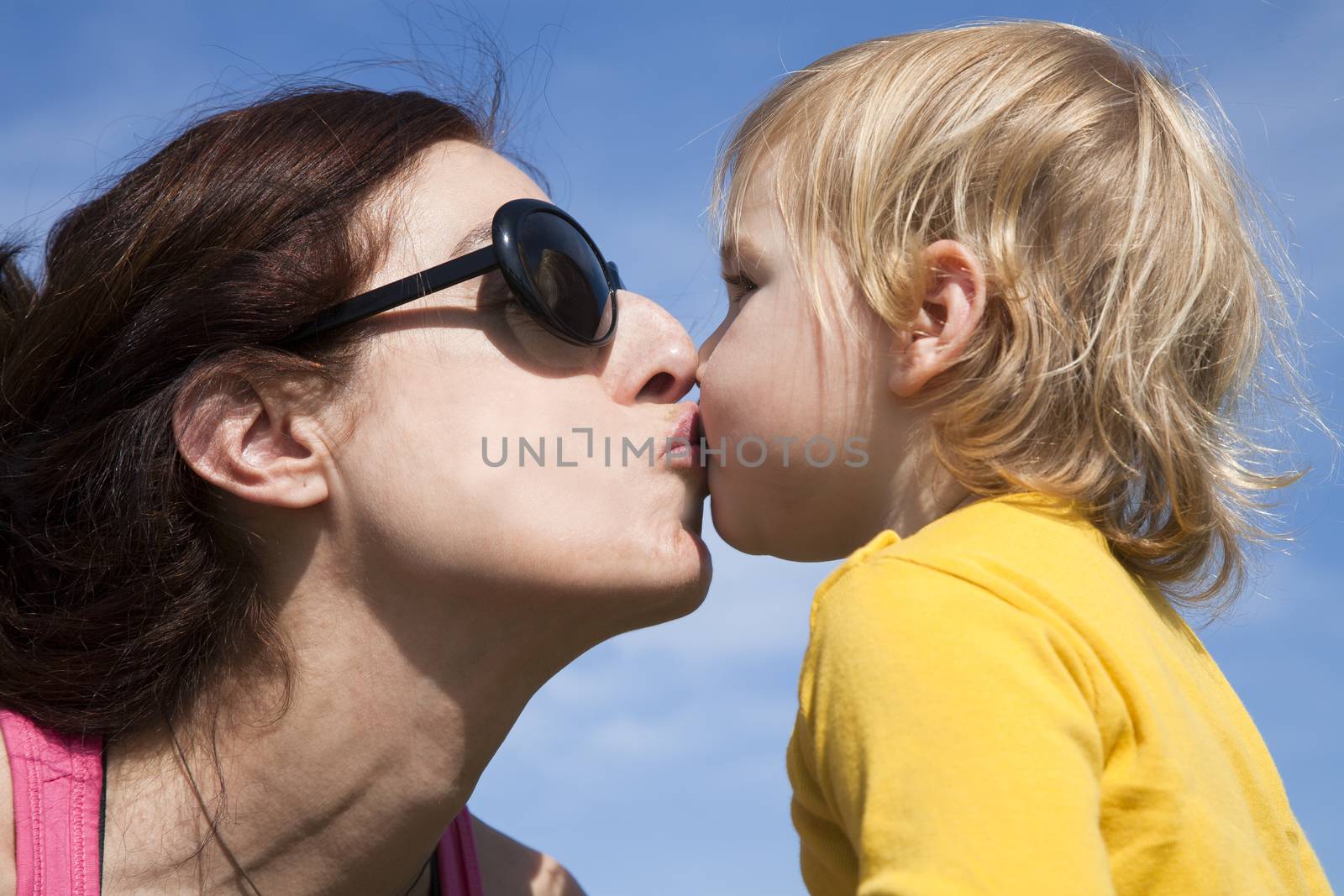 two years old age blonde baby yellow shirt kissing in mouth to brunette woman mother sunglasses with blue sky background