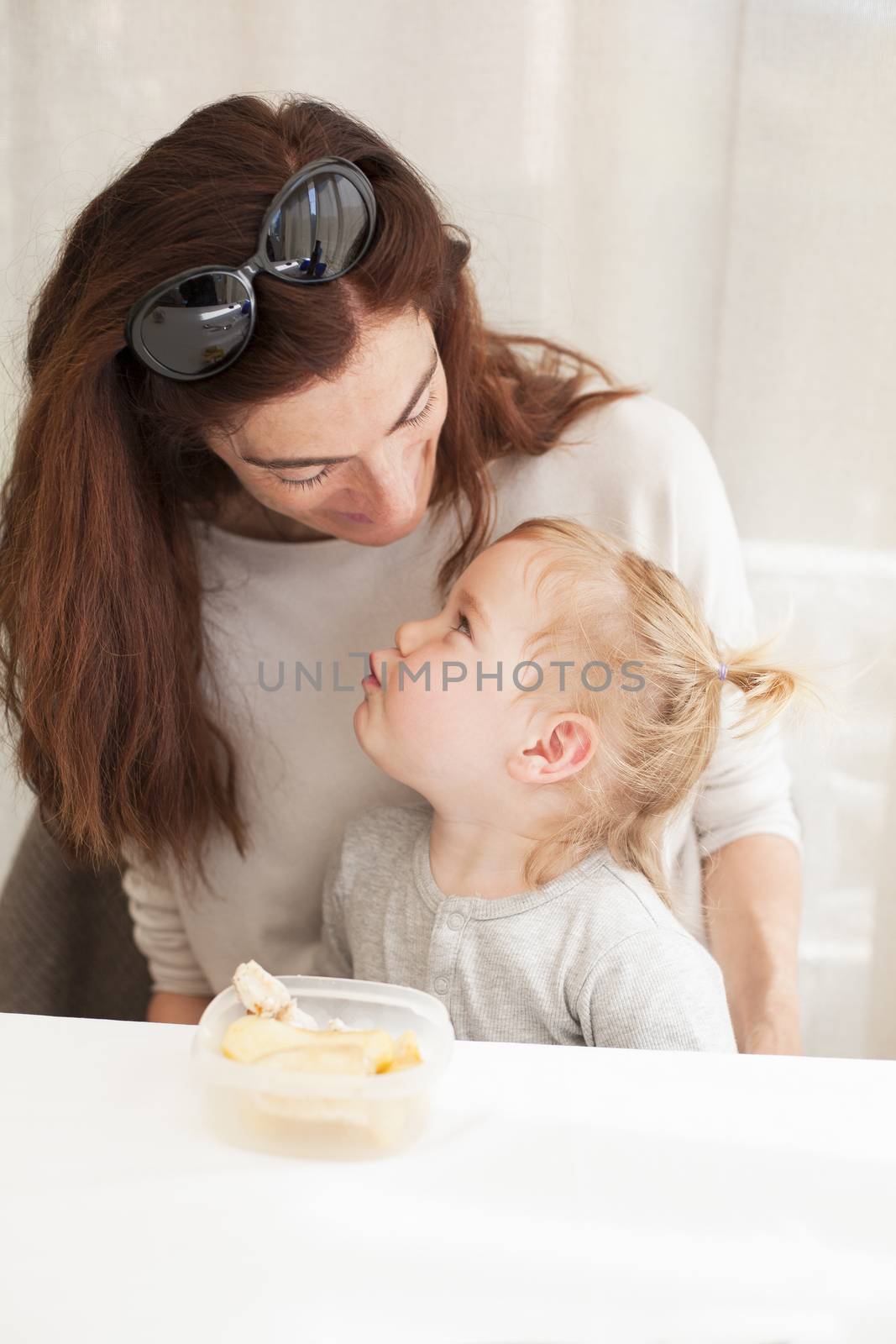 portrait of two years age blonde baby with grey shirt eating chicken from tupperware sitting on legs of mother woman in white table