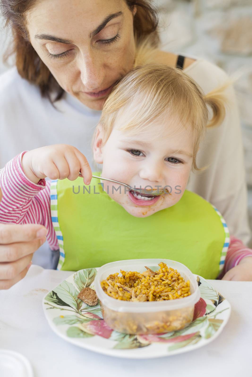 portrait of two years age blonde baby with green bib eating rice paella from tupperware sitting on legs of mother woman in white table