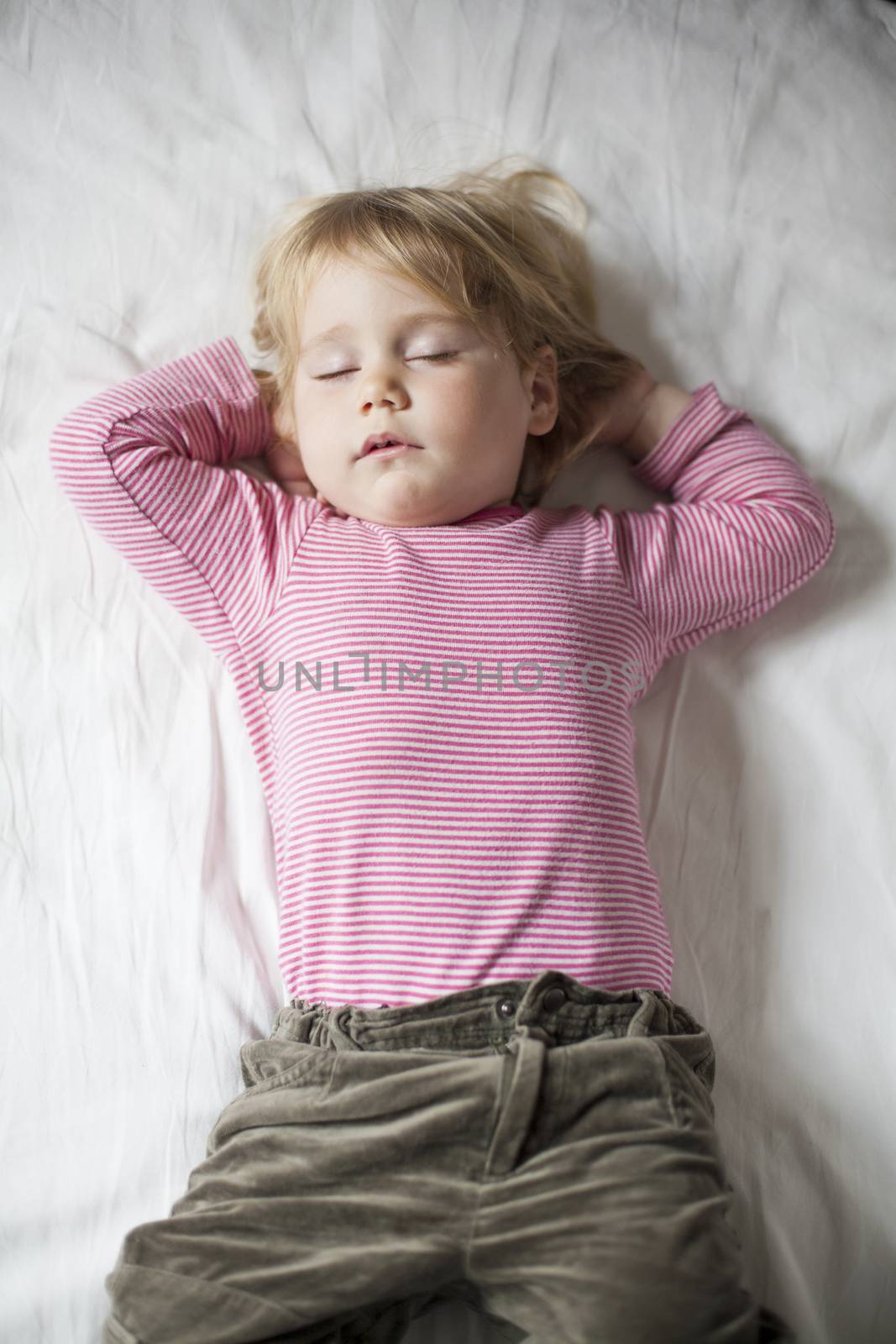blonde caucasian baby face two years old age with red striped shirt sleeping on white sheets with hands down her head