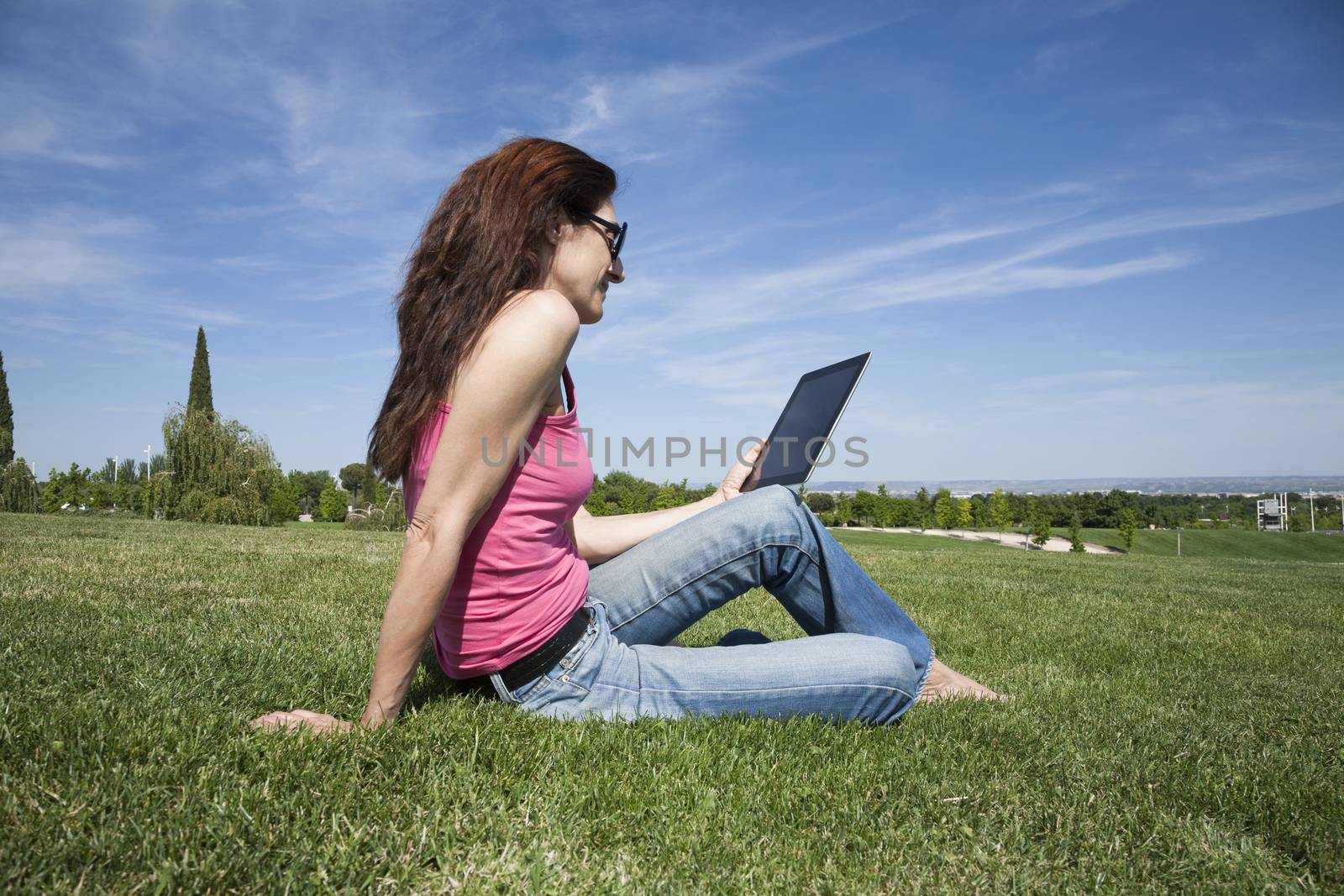 redhead woman blue jeans trousers pink shirt reading digital tablet blank screen sitting on green grass lawn in park
