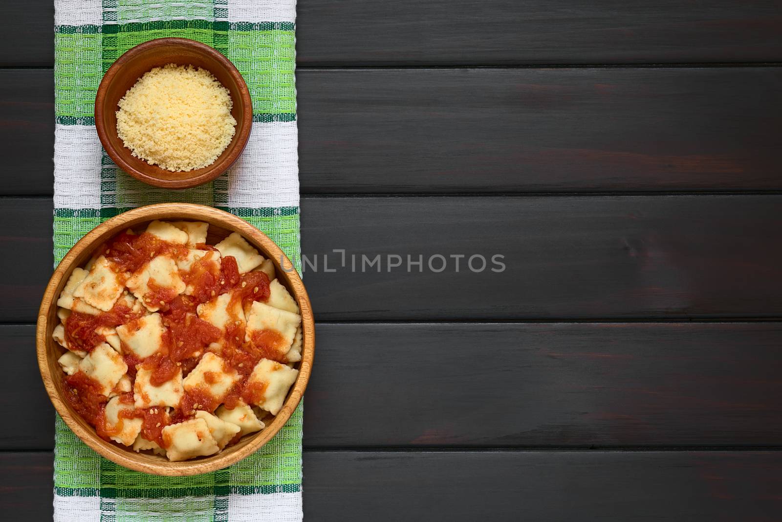 Cooked ravioli with homemade tomato sauce in wooden bowl with grated cheese in small bowl, photographed overhead on dark wood with natural light