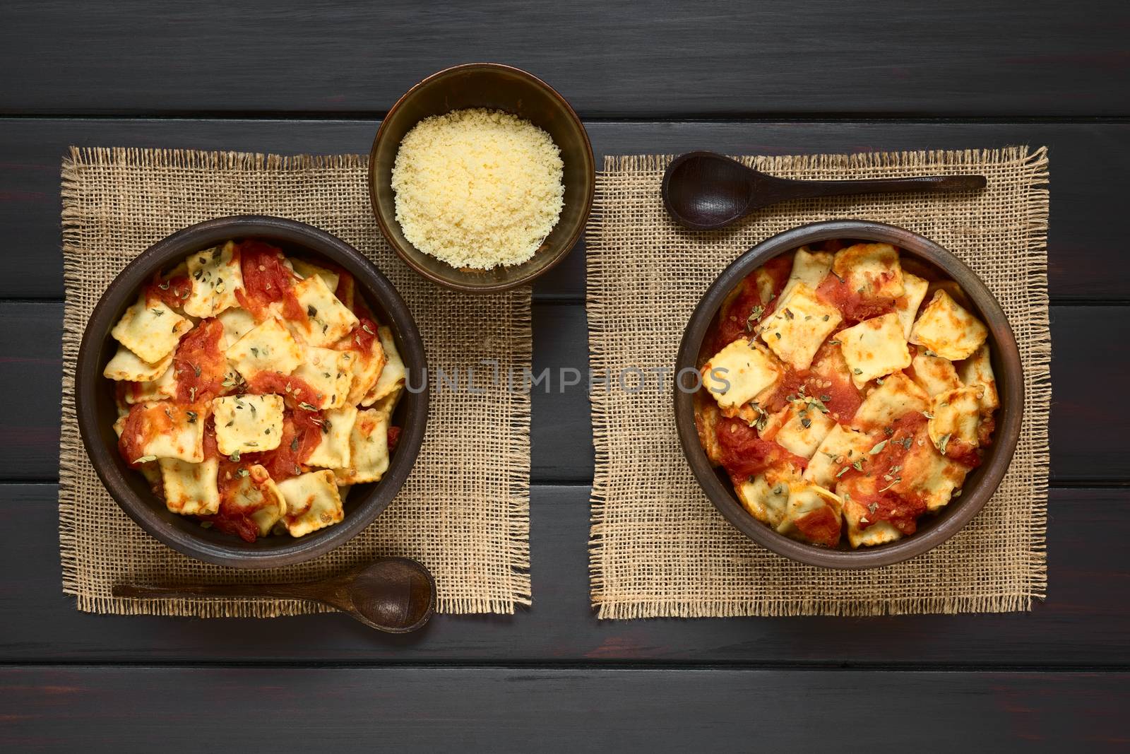 Baked ravioli with homemade tomato sauce in rustic bowls with grated cheese and wooden spoons on the side, photographed overhead on dark wood with natural light