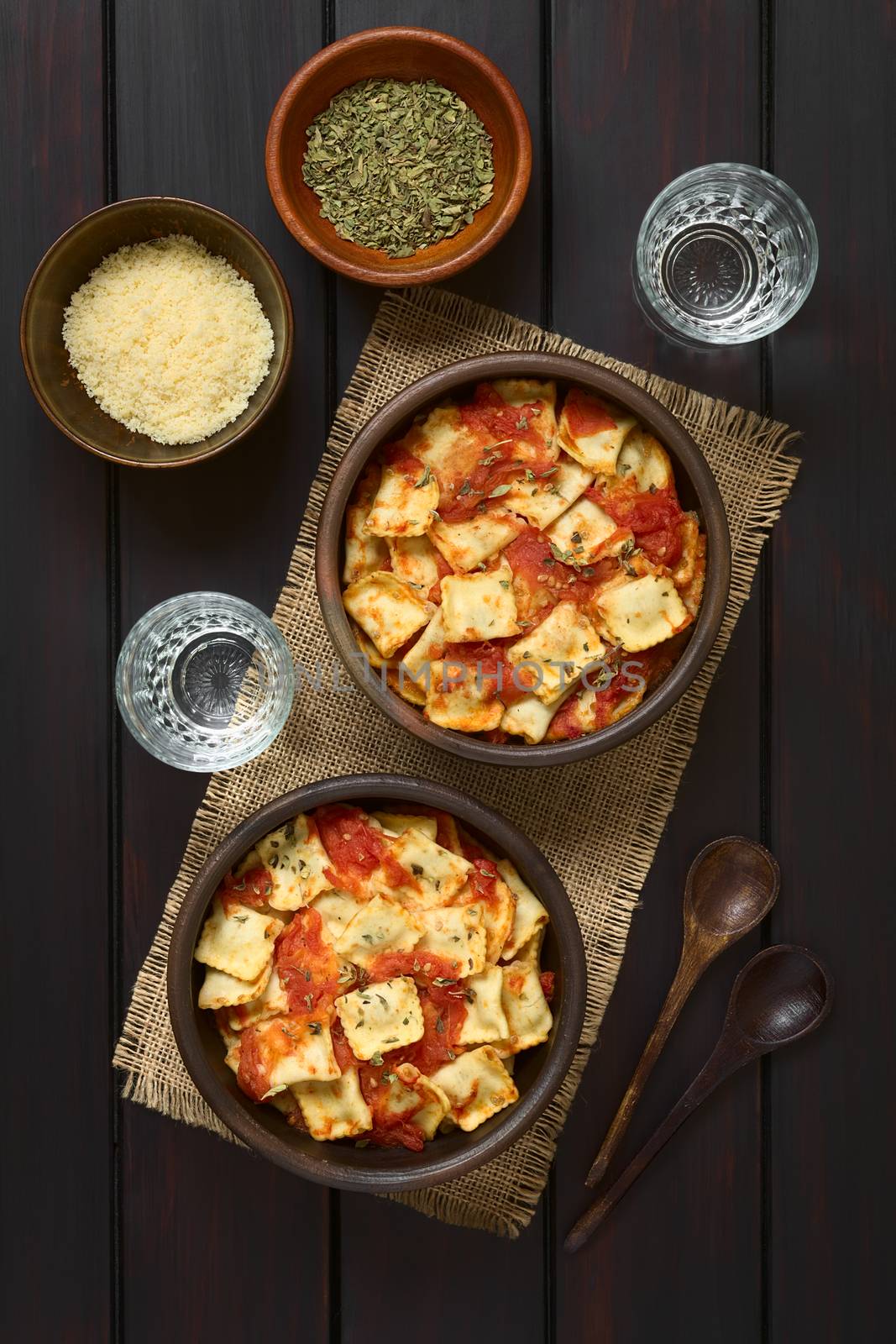 Baked ravioli with homemade tomato sauce in rustic bowls with grated cheese and dried oregano in small bowls, glasses of water and wooden spoons on the side, photographed overhead on dark wood with natural light