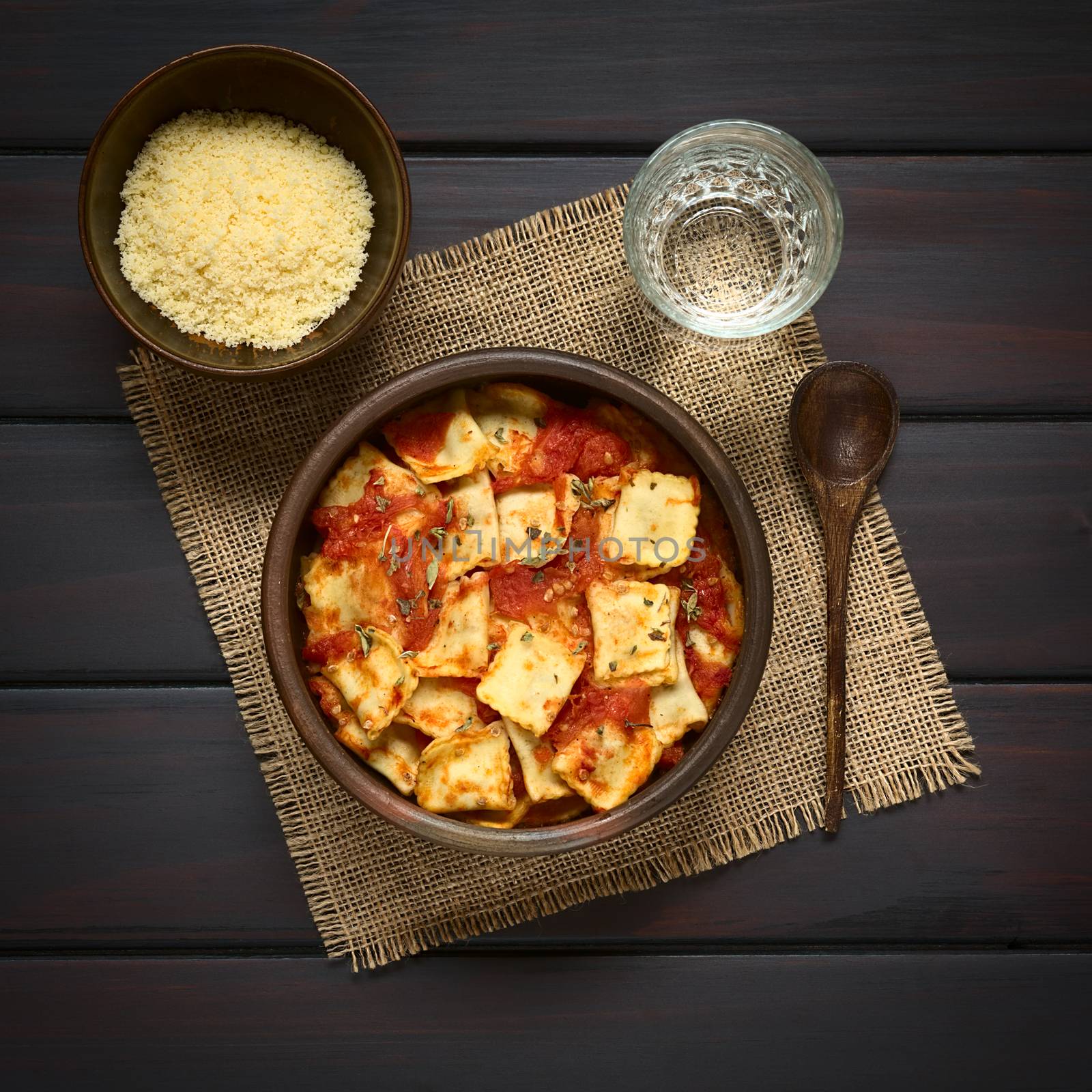 Baked ravioli with homemade tomato sauce in rustic bowl with grated cheese in small bowl, glass of water and wooden spoon on the side, photographed overhead on dark wood with natural light