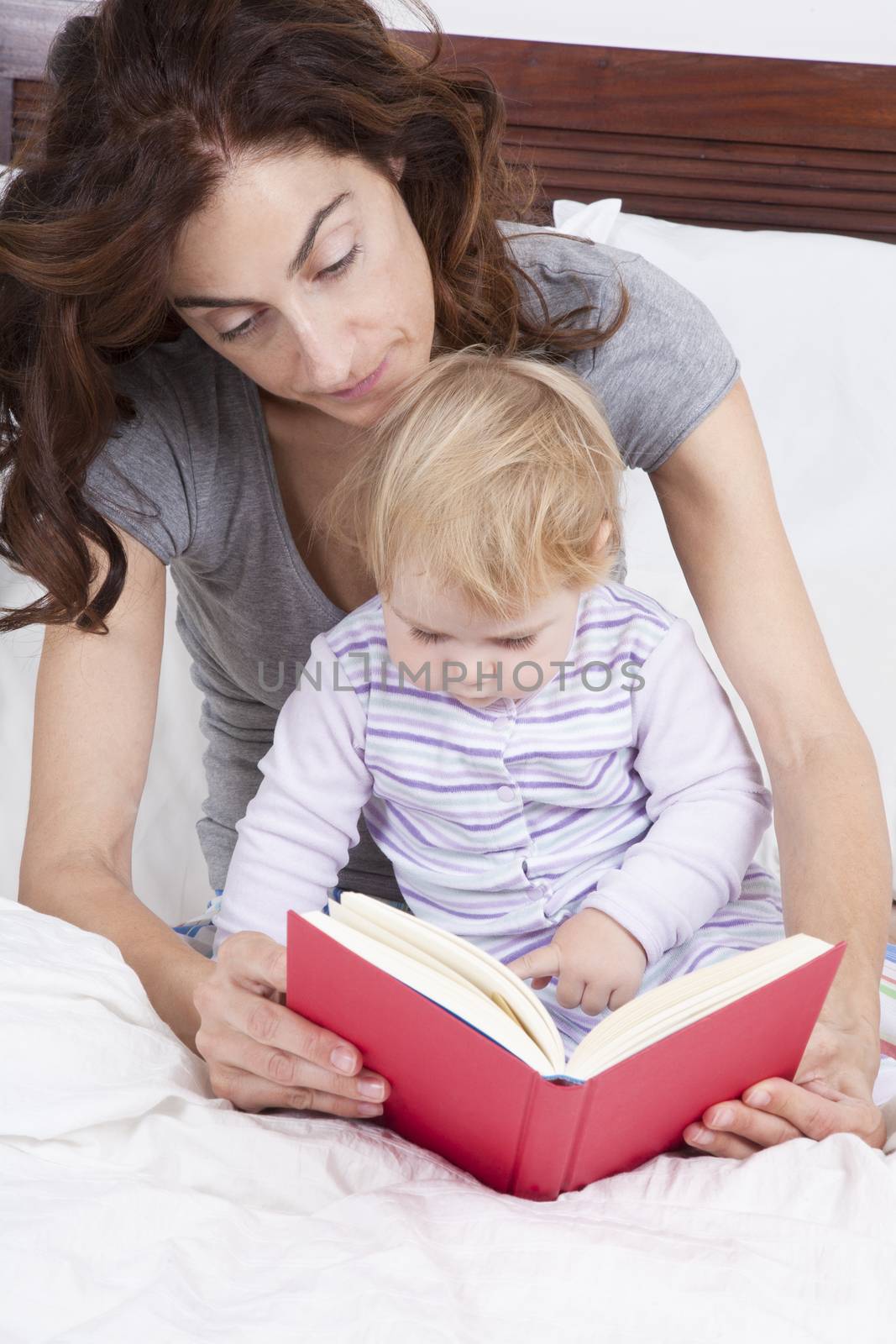 eighteen month aged blonde baby with brunette woman mother reading tale story red book in white bed