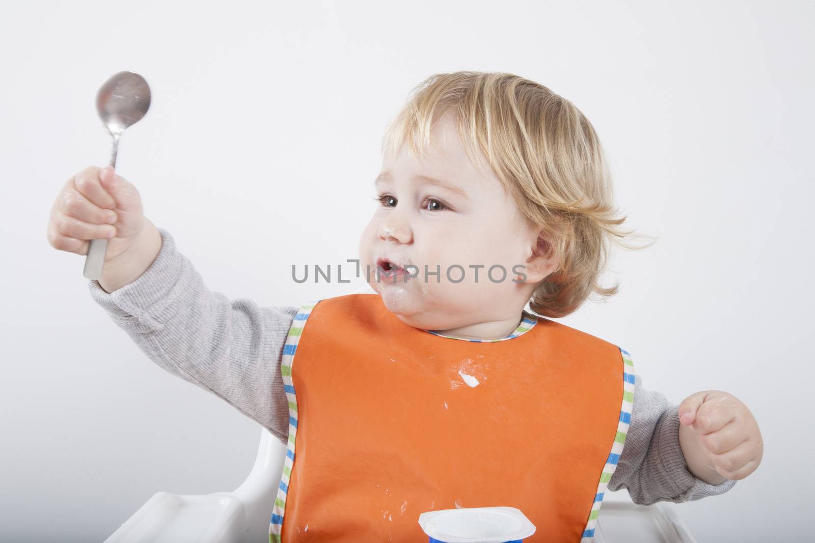 blonde caucasian baby seventeen month age orange bib grey sweater on white high chair with spoon hand up