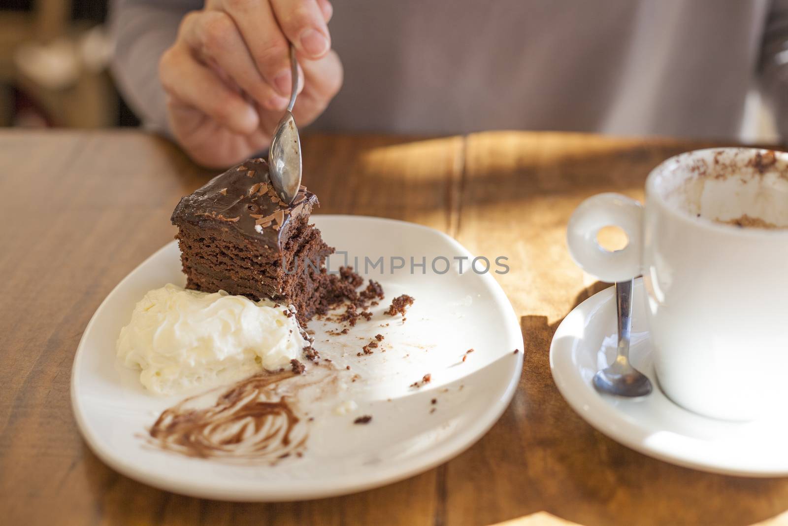 woman hands taking chocolate cake piece with spoon and white small cup cappuccino coffee on light brown wooden table