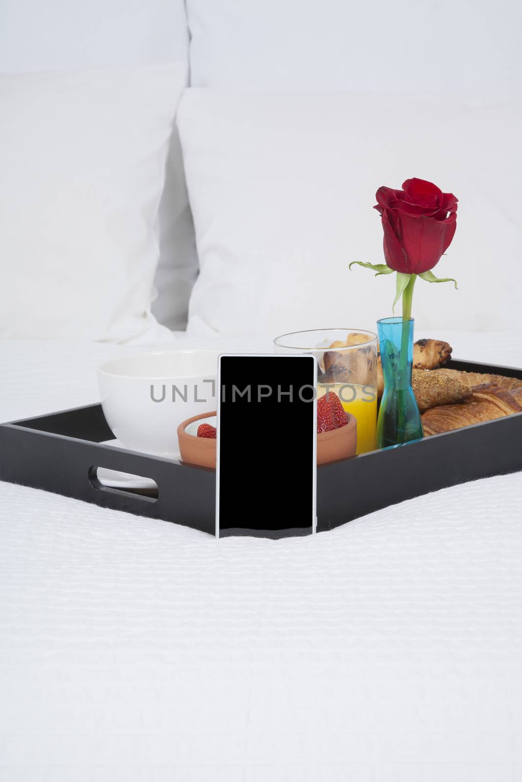 white quilt bed breakfast black tray croissants orange juice strawberry kiwi cupcake red rose flower and mobile phone blank screen