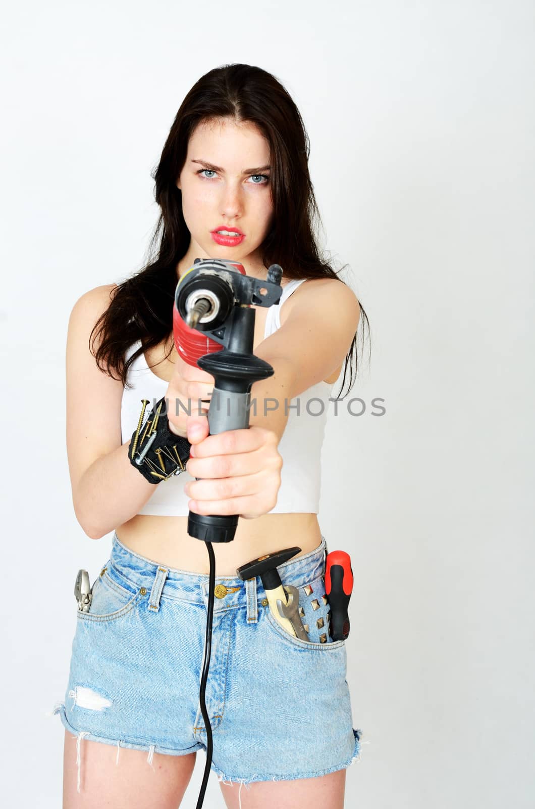 Young female model wearing white top and jeans' shorts. Girl holding big driller, keeping key, screwdriver and small hammer in her pocket.