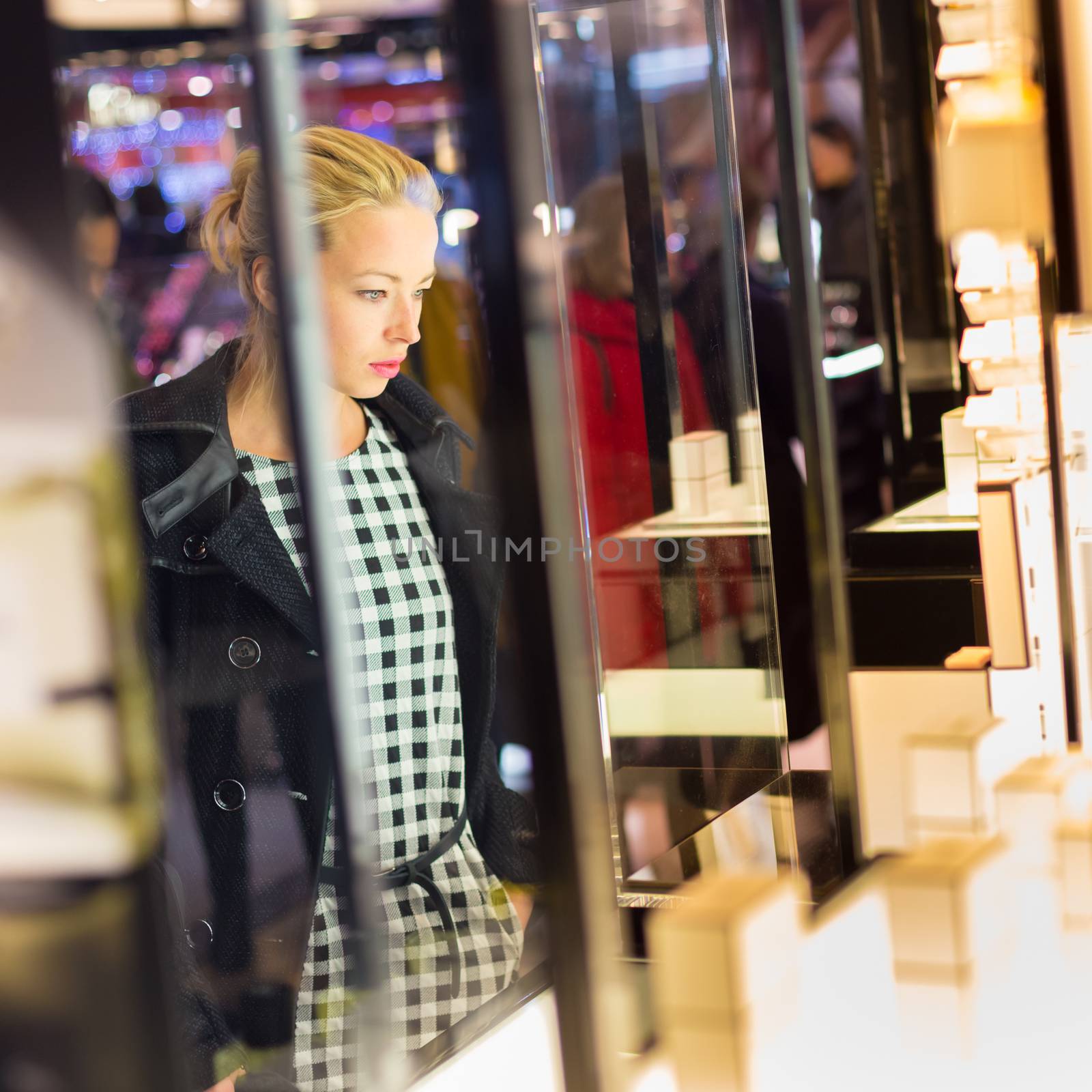 Beautiful blonde lady standing in front of showcase in beauty store, admiring new perfume collection.