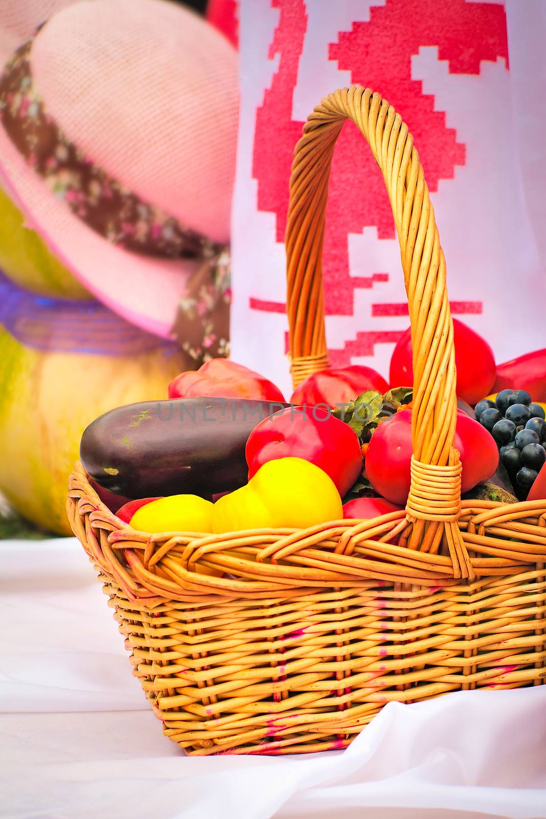 On a white towel there is a beautiful basket with tomatoes, eggplants, pepper, grapes for sale at fair.