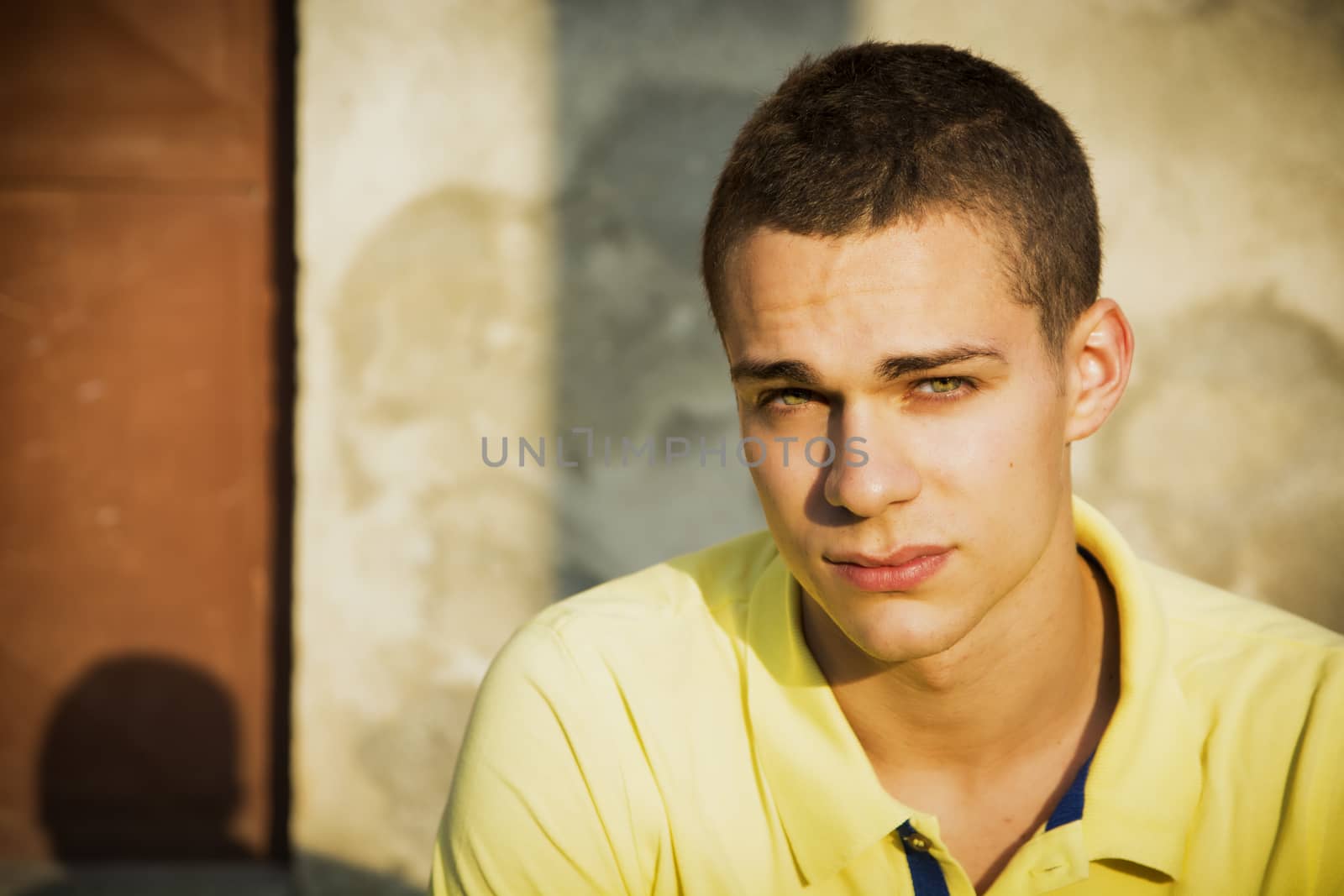 Attractive green eyed young man leaning against rough concrete wall, looking at camera
