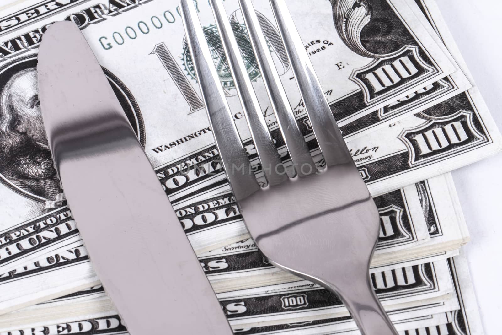 Close up detailed view of one hundred dollar banknotes under a steel fork and knife, isolated on white background.
