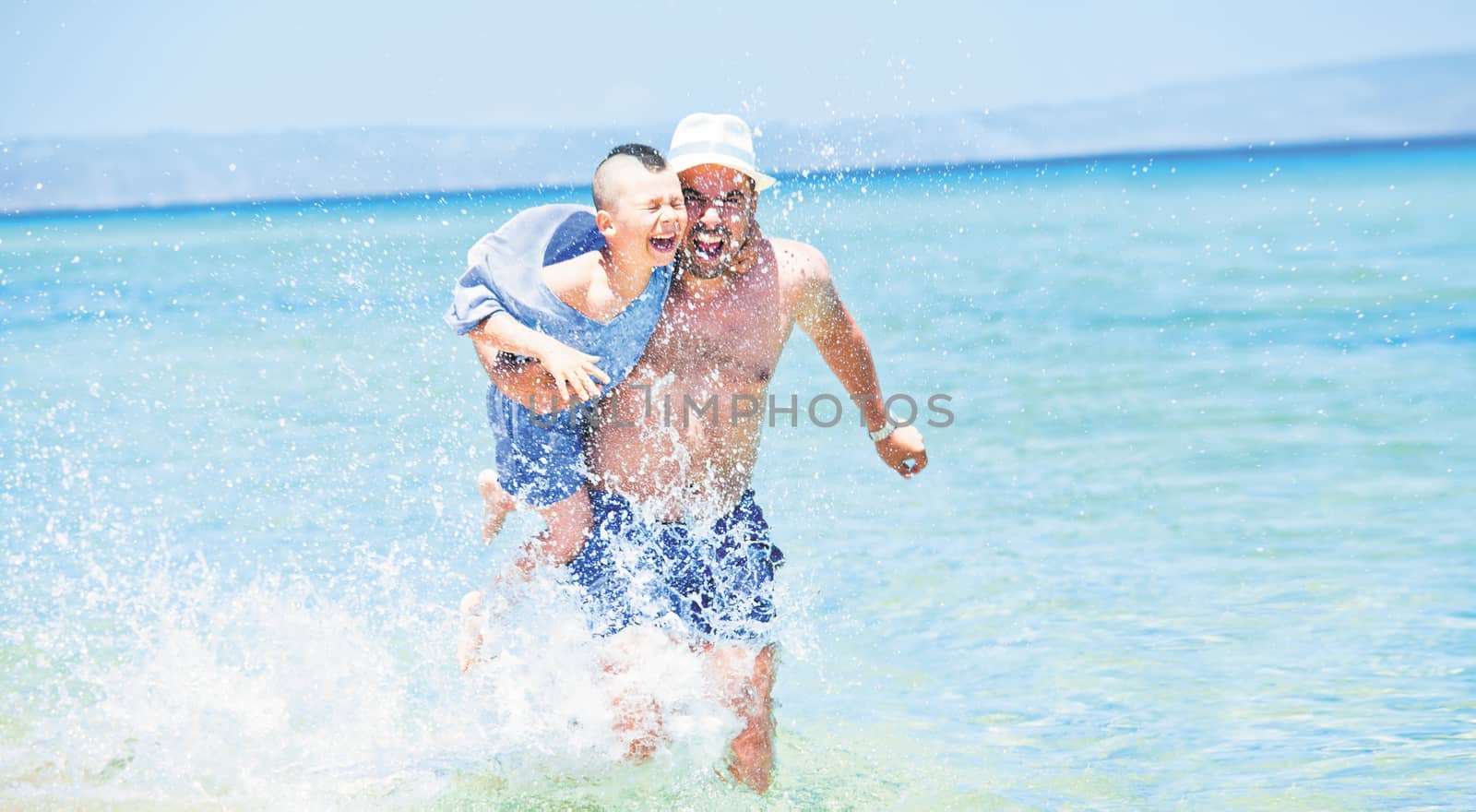 Father holding his son is running through sea or ocean, splashing water arround.