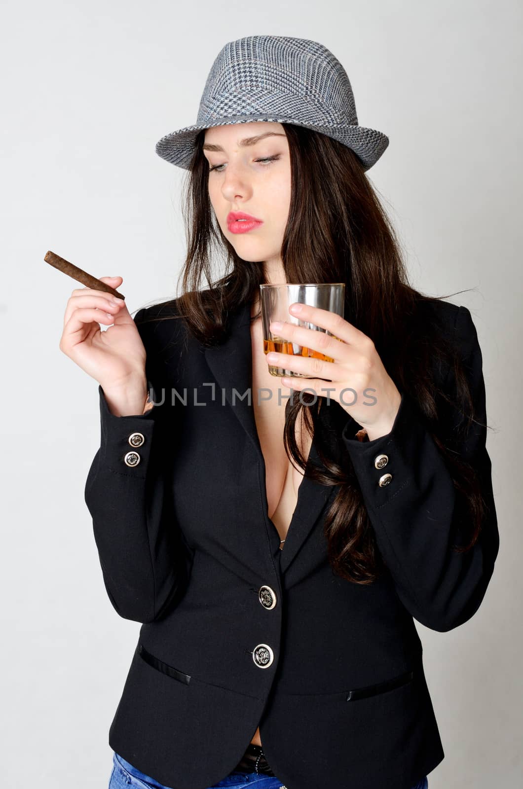 Young female model holds cigar and glass of whiksy. Girl puts black tail-coat and grey hat.
