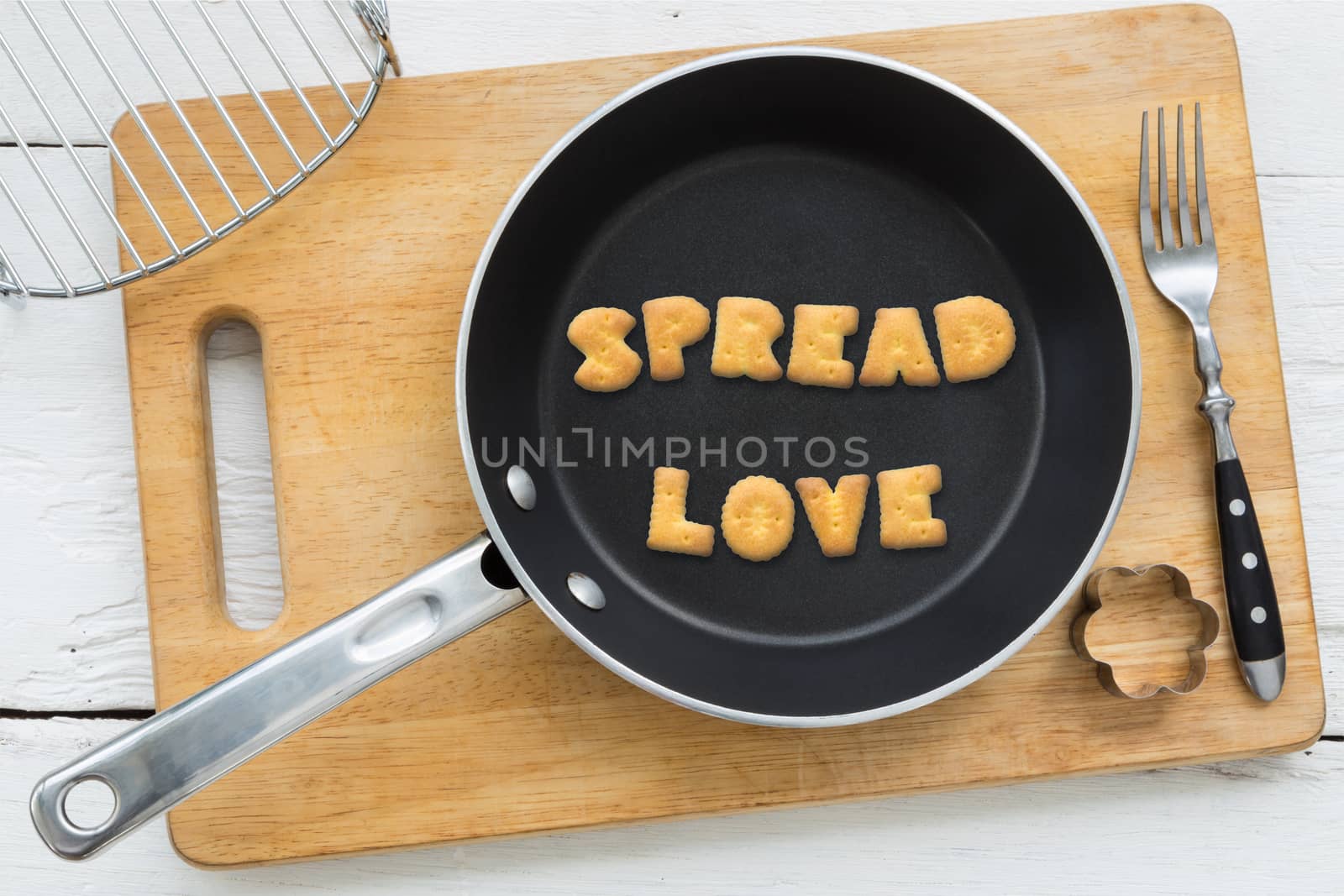 Top view of letter collage made of biscuits. Word SPREAD LOVE putting in black frying pan. Other cooking equipments: fork, cookie cutter and chopping board putting on white wooden table, vintage style image.