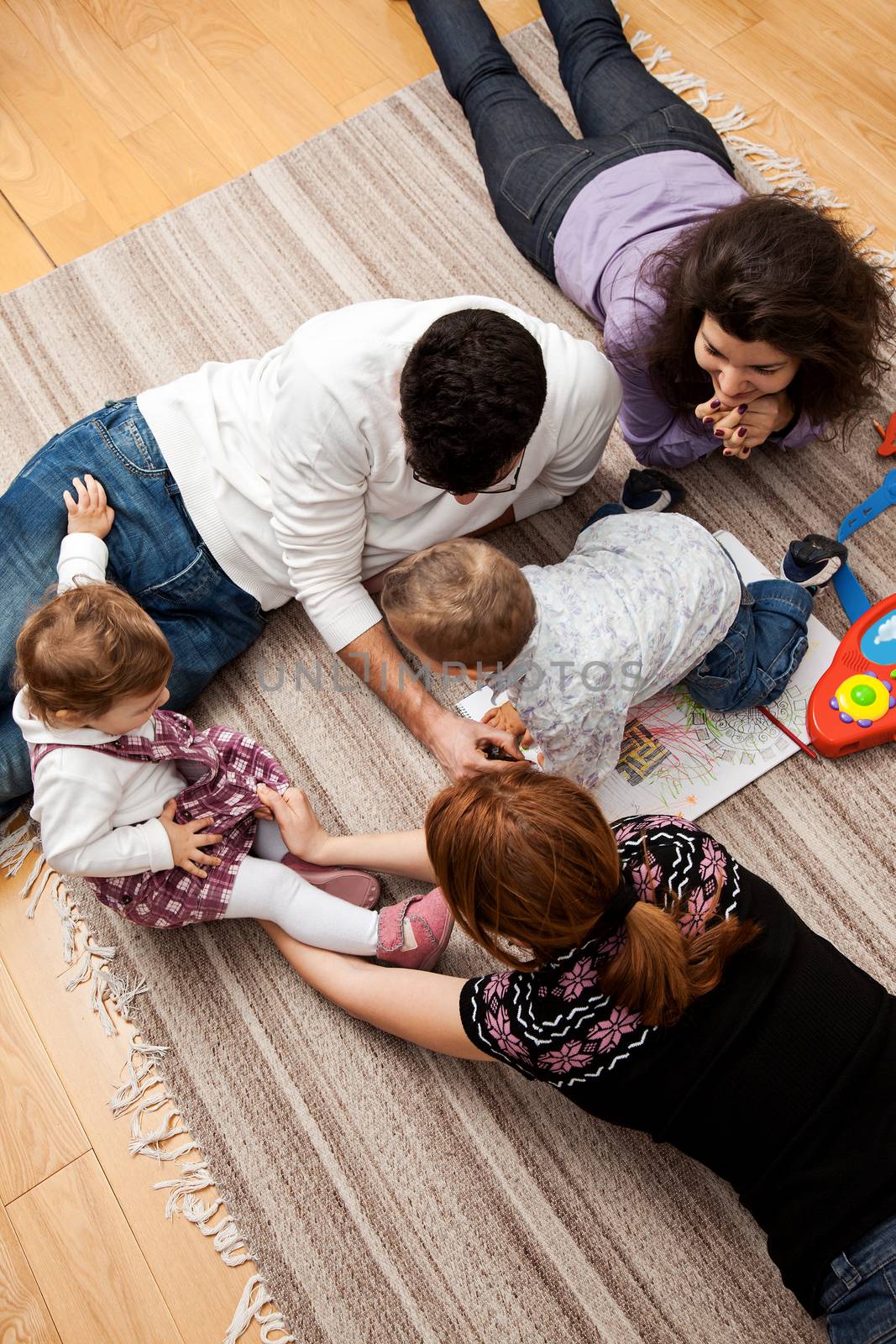 family group of five - two babies and three adults lying on the carpet, playing together.