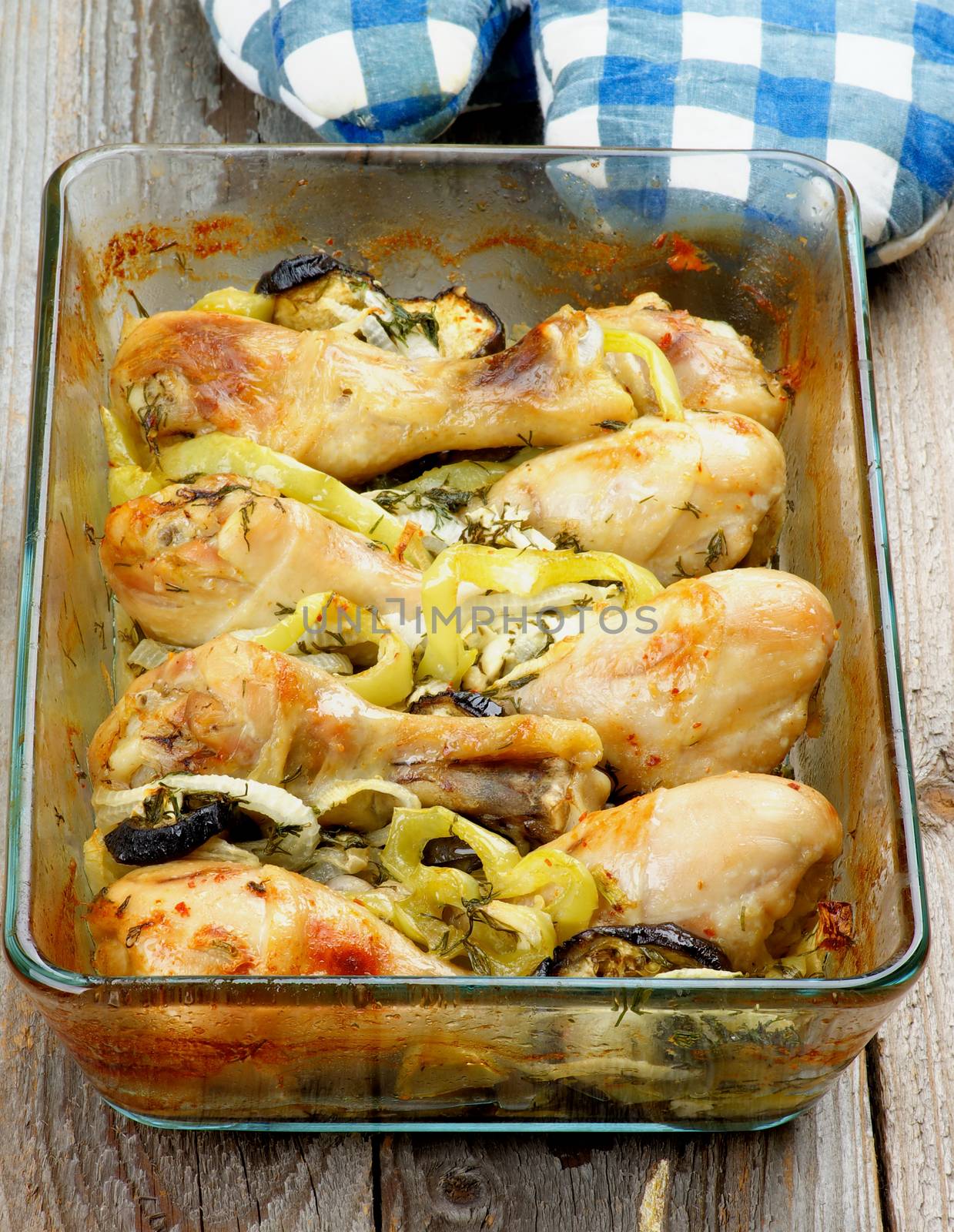 Delicious Baked Chicken Legs with Vegetables in Glass Casserole with Checkered Oven-Gloves closeup on Rustic Wooden background
